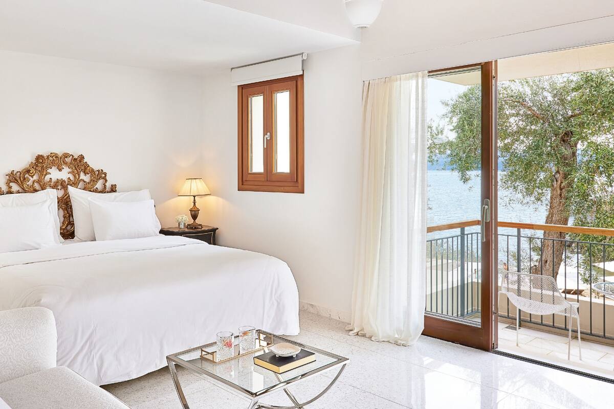 A hotel room with a bed, a nightstand, a glass coffee table, and a balcony with a view of the sea.