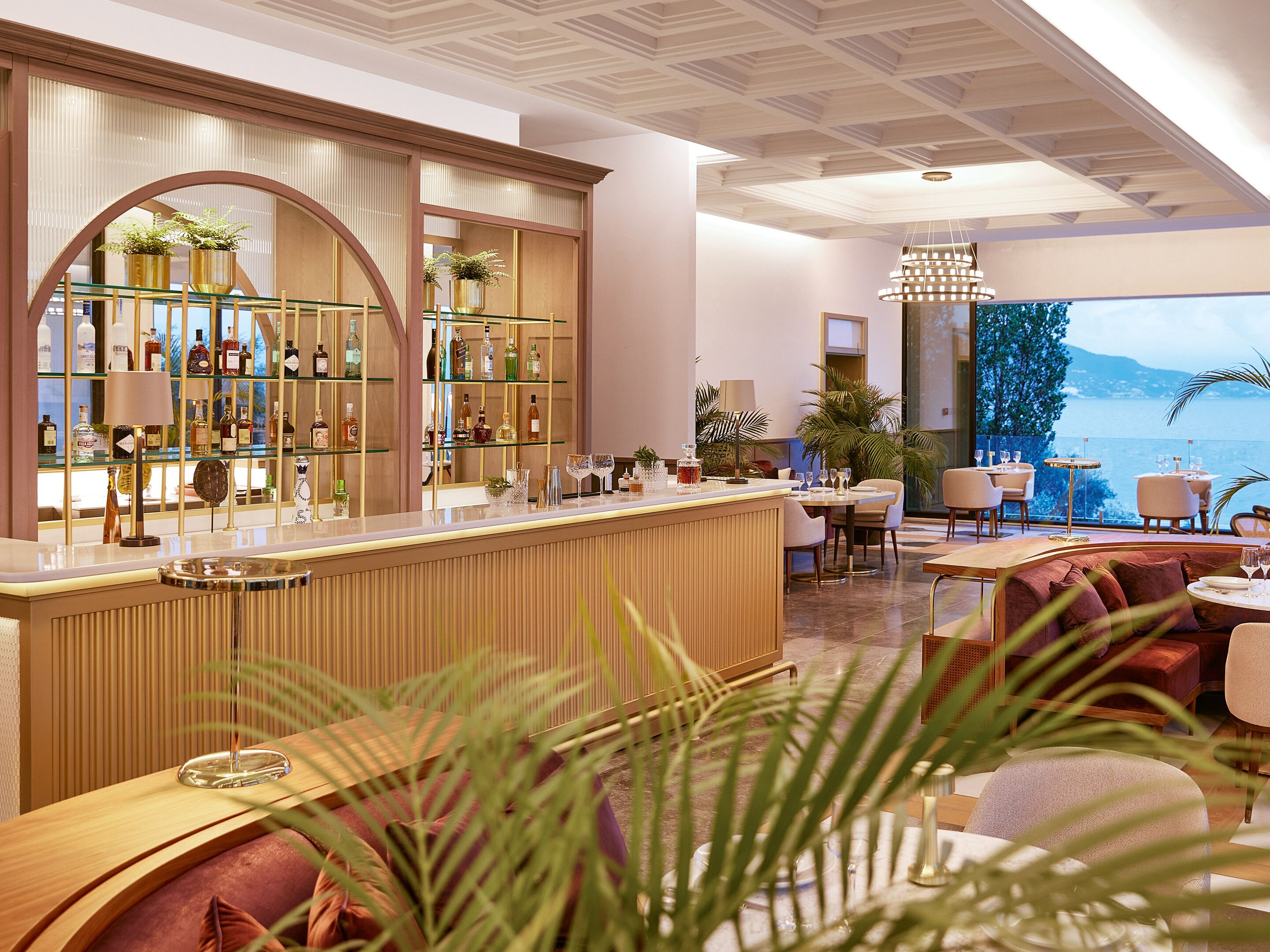 A bar area with a wooden counter, shelves with bottles, and a view of the sea.