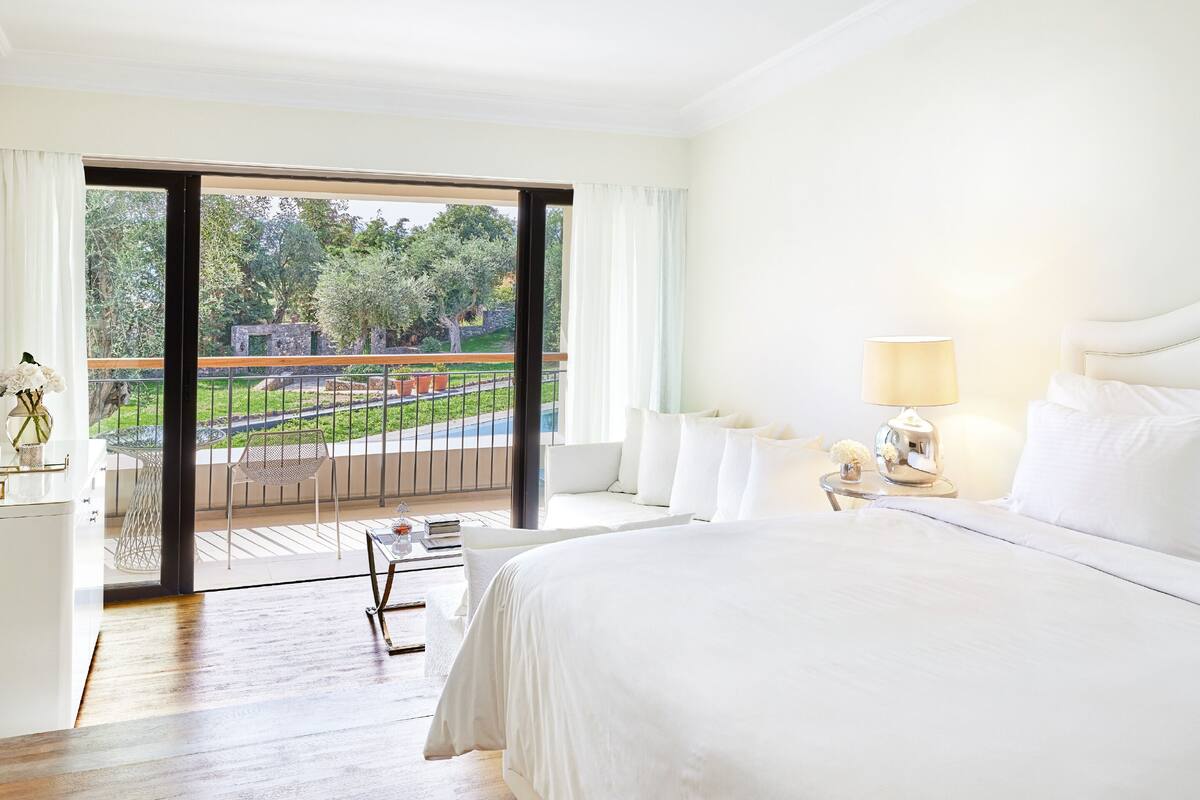 A hotel room with a large bed, a bedside table with a lamp, a view of a garden through a sliding door, and a small table with a vase and a book.