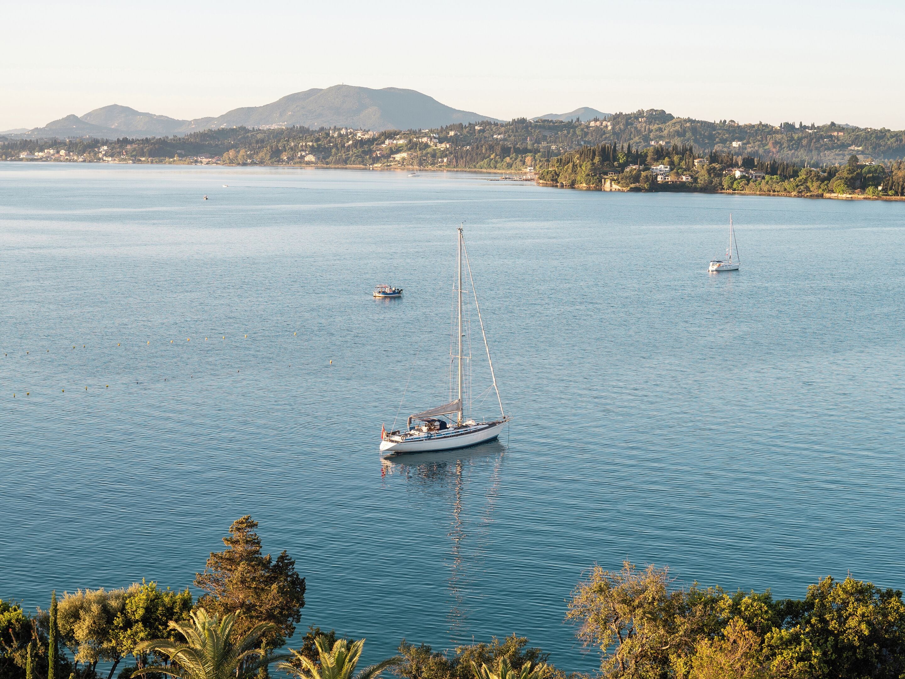 A calm bay with several sailboats, a distant shoreline with buildings, and a mountain in the background.