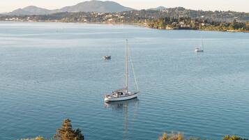 A calm bay with several sailboats, a distant shoreline with buildings, and a mountain in the background.