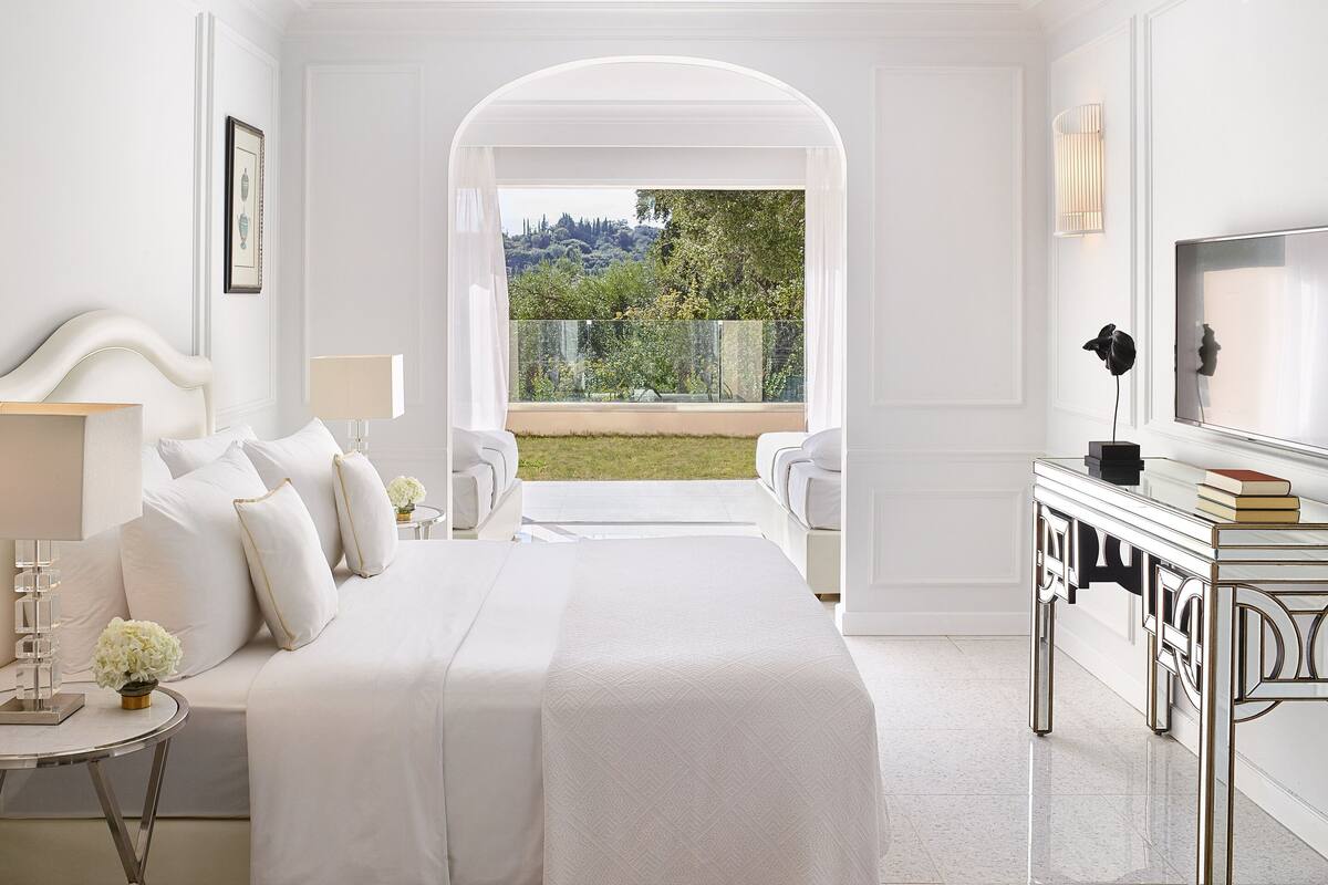 A spacious bedroom with a large bed, a bedside table with a lamp, a TV, and a view of a garden.
