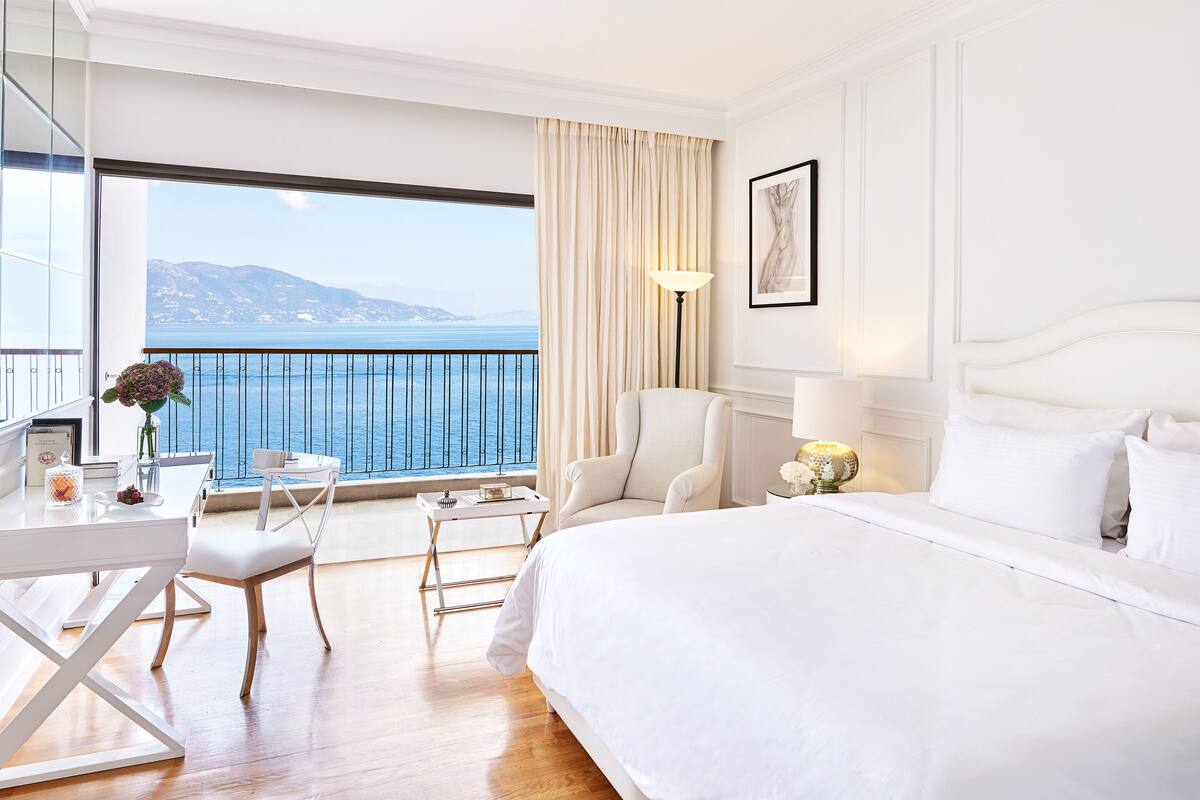 A hotel room with a large bed, a desk with a chair, a small table, and a view of the sea.