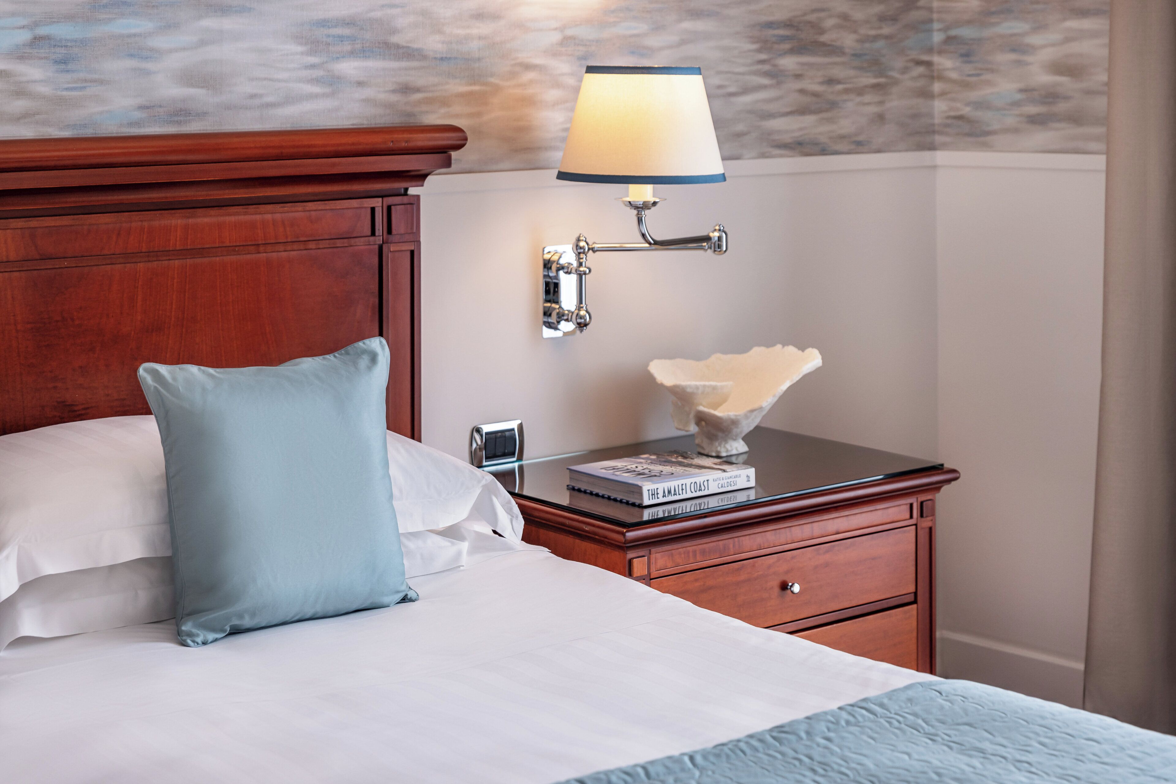 Una cama bien hecha con un almoháron azul claro, una mesita de noche de madera con una lámpara y un libro titulado 'The Amalfi Coast'.