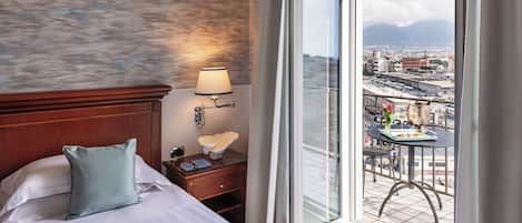 A hotel room with a large bed, a nightstand, a lamp, and a view of the city through a balcony.