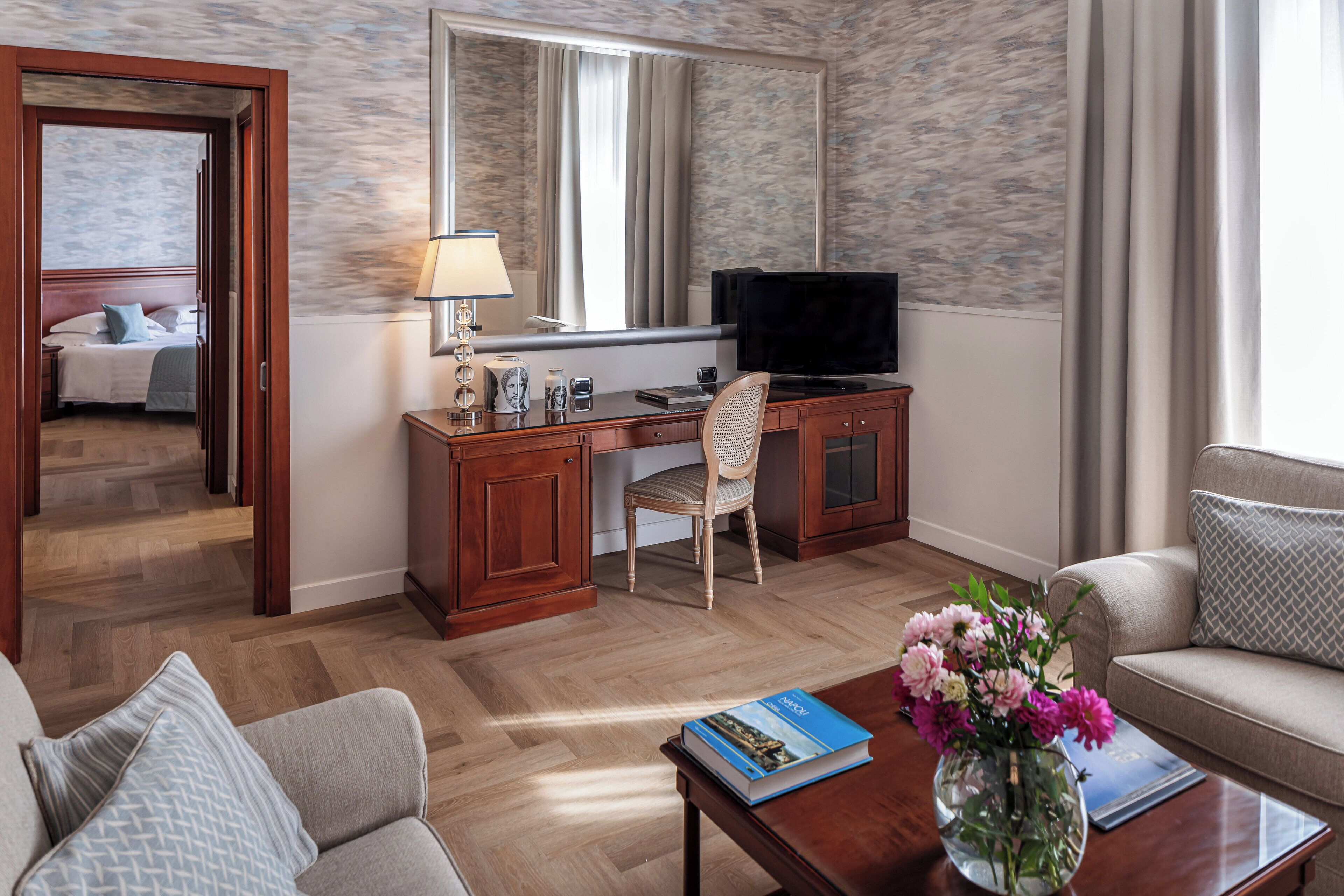 A hotel room with a wooden desk, a television, a chair, a sofa, and a coffee table with a book and flowers.