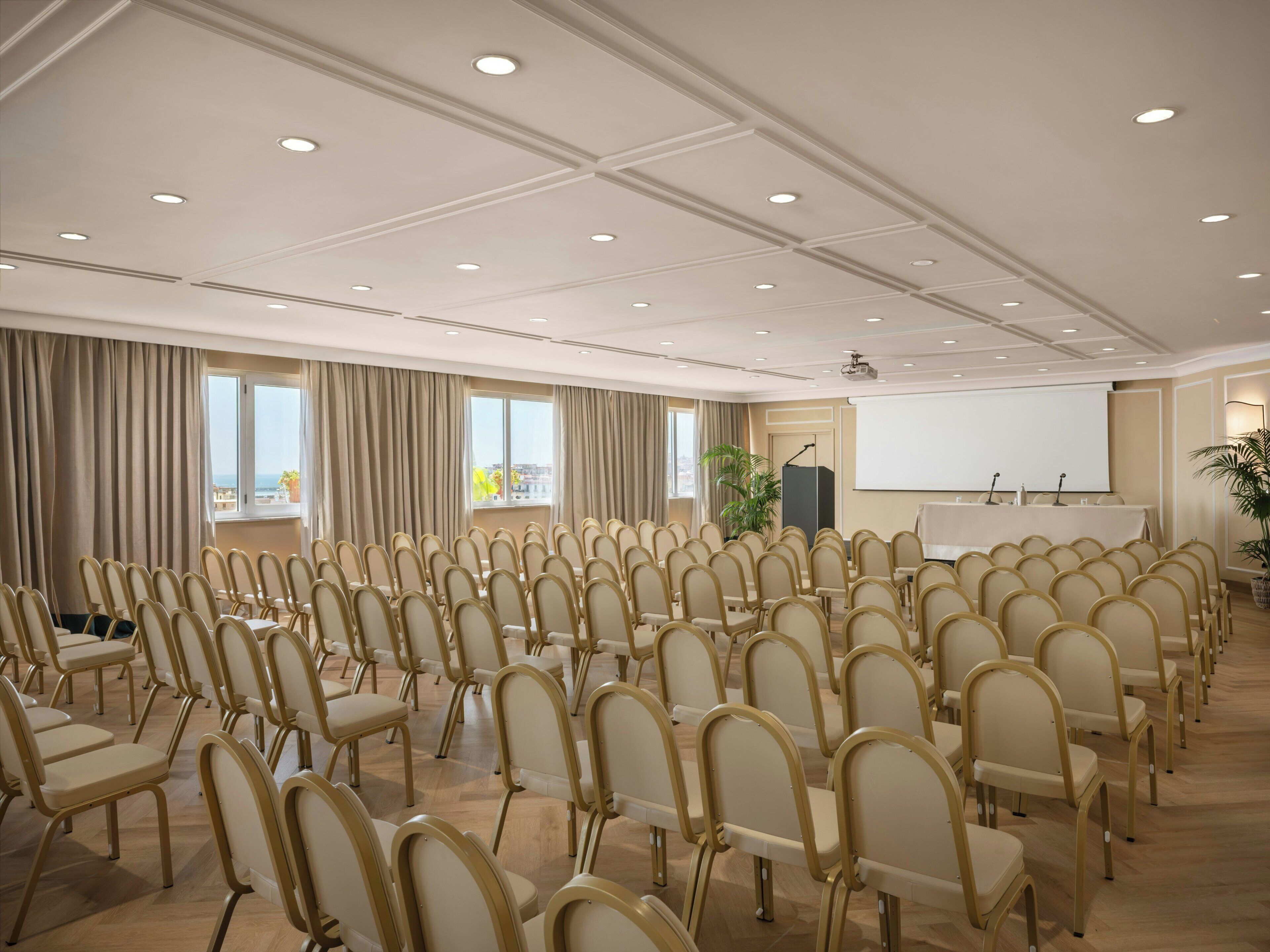 A conference room with rows of chairs, a projector screen, and a large window with curtains.