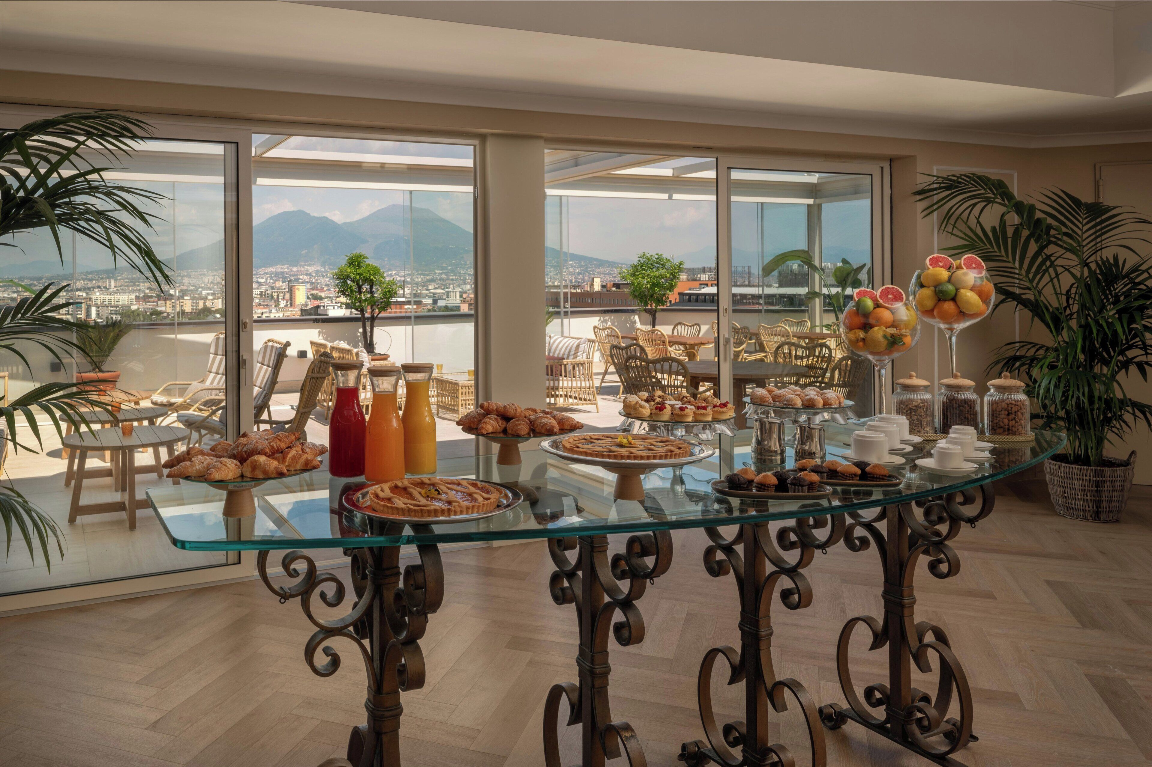 Un buffet de petit-déjeuner proposant des viennoiseries, des fruits et diverses boissons, le tout disposé sur une table en verre dans une pièce dotée de grandes fenêtres donnant sur un panorama urbain.