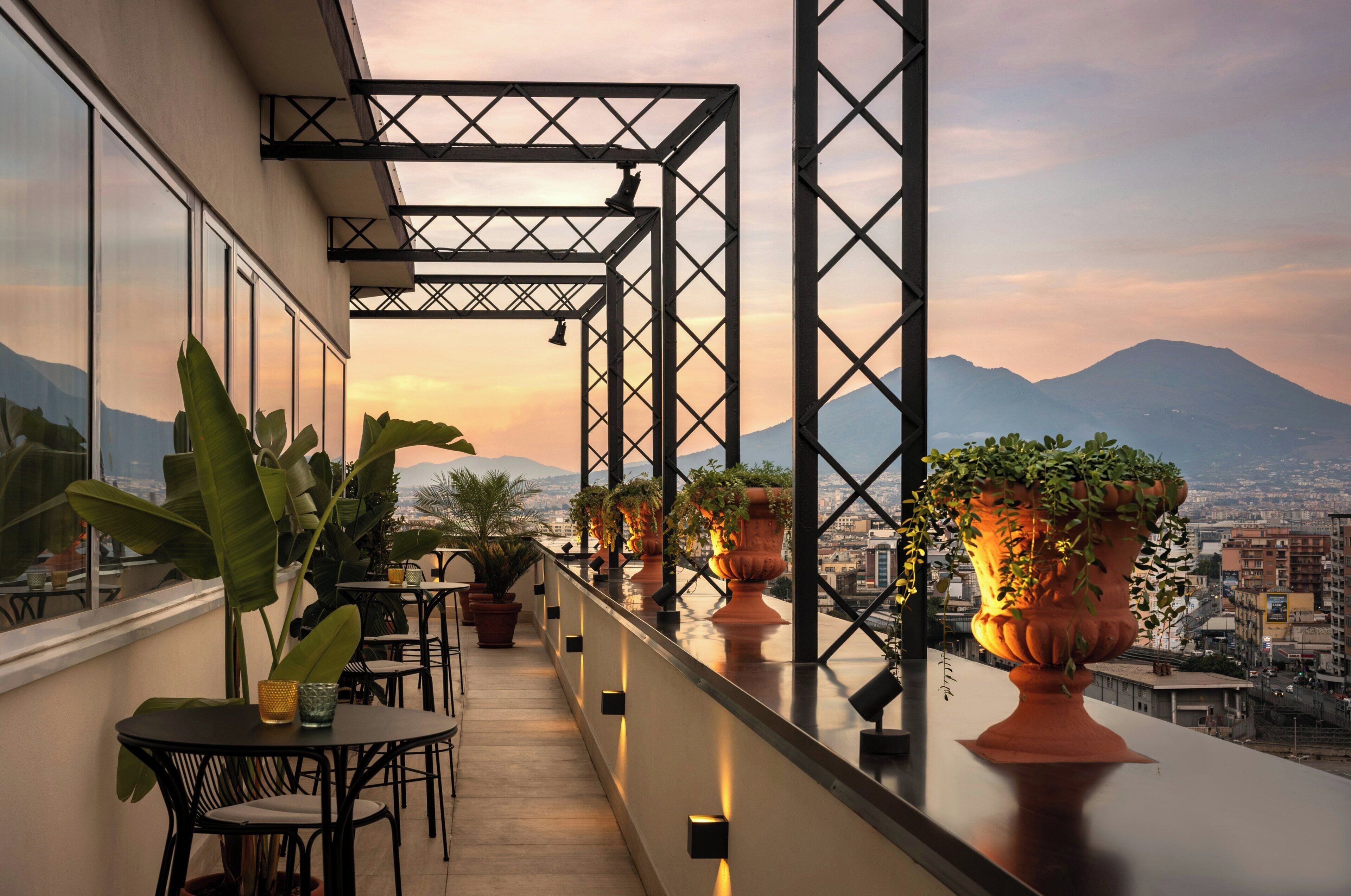 A balcony with potted plants, a table, and chairs, overlooking a cityscape with mountains in the distance.