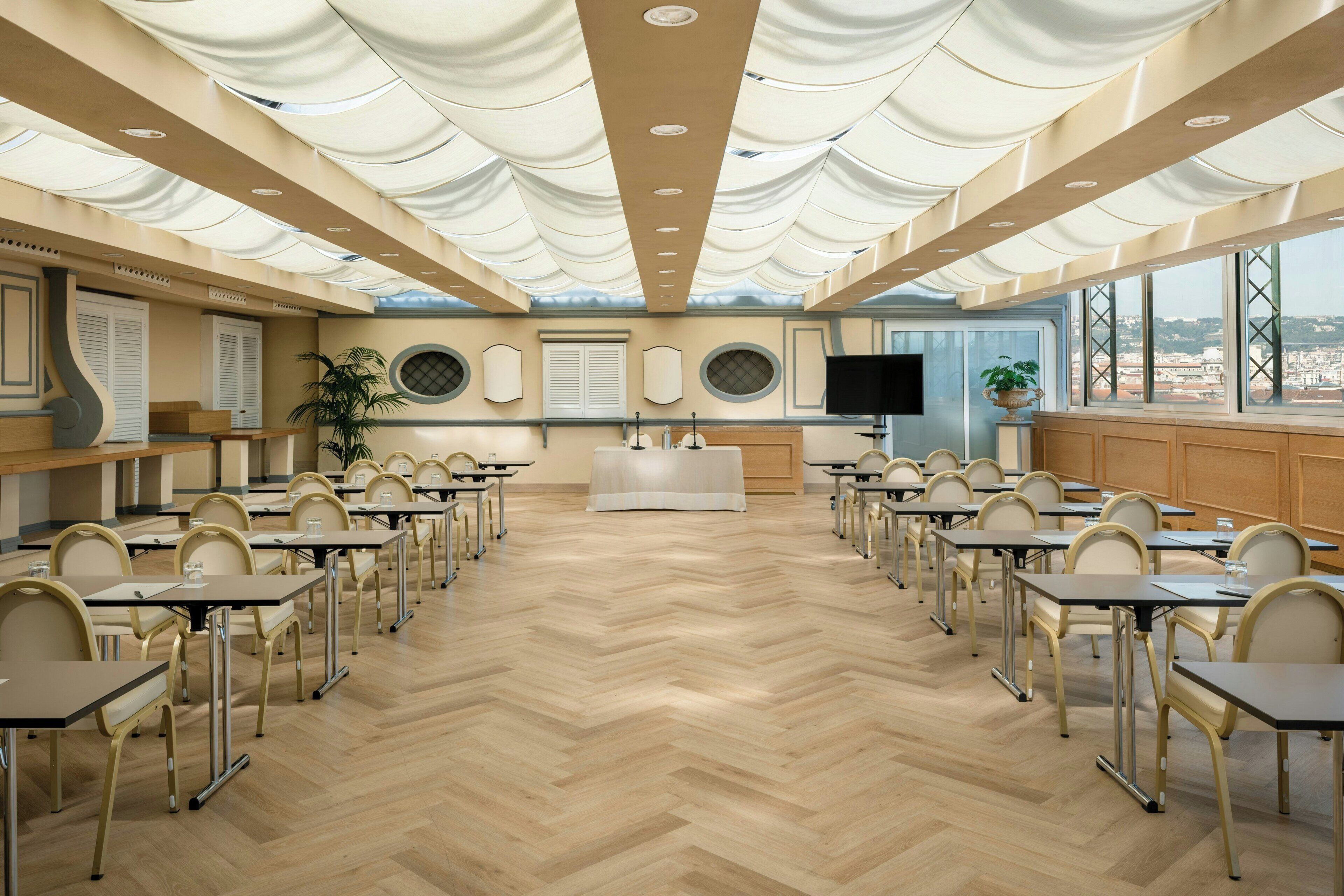 A conference room with a herringbone floor, multiple tables, chairs, a large screen, and a view through the windows.