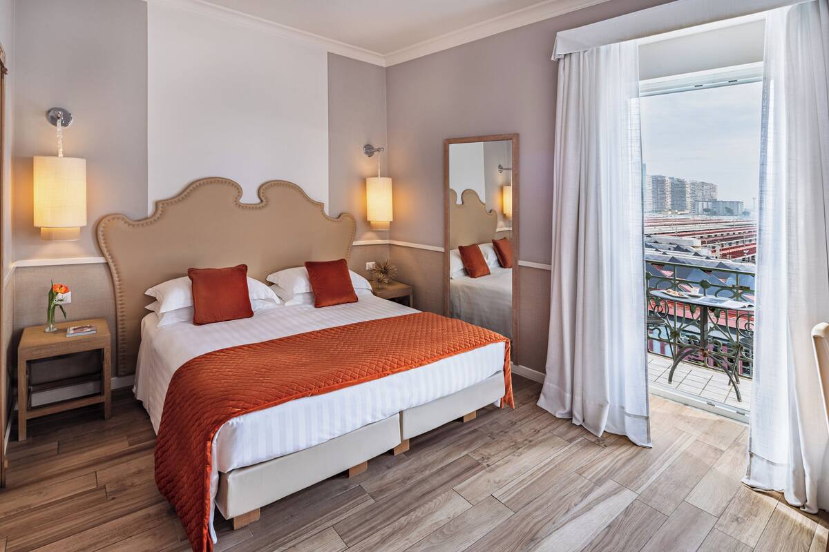 A hotel room with a large bed, two bedside tables, a nightstand, a mirror, and a view of a cityscape through a balcony.