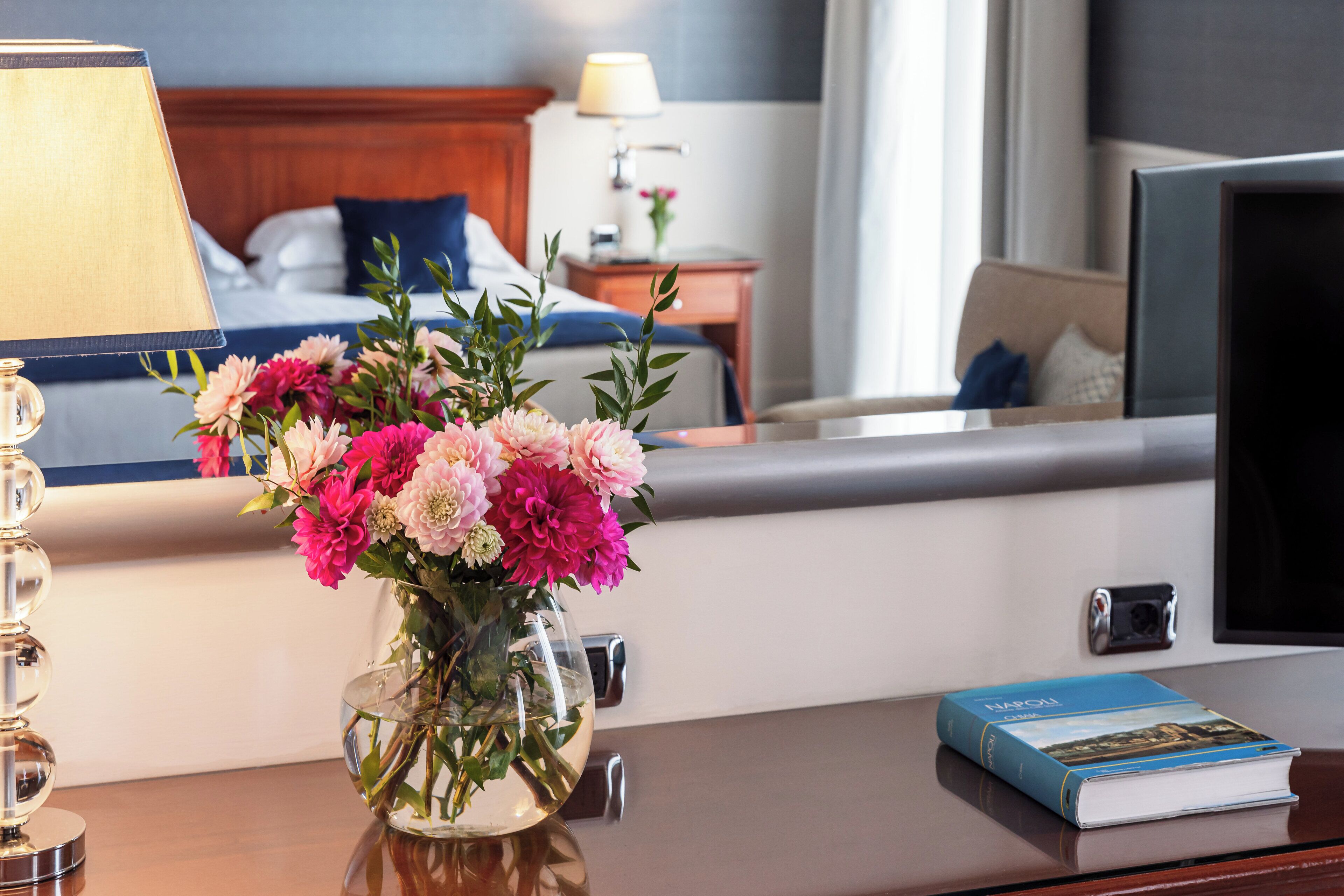 A hotel room with a bed, a bedside table with a lamp, a vase of flowers, and a book on the table.