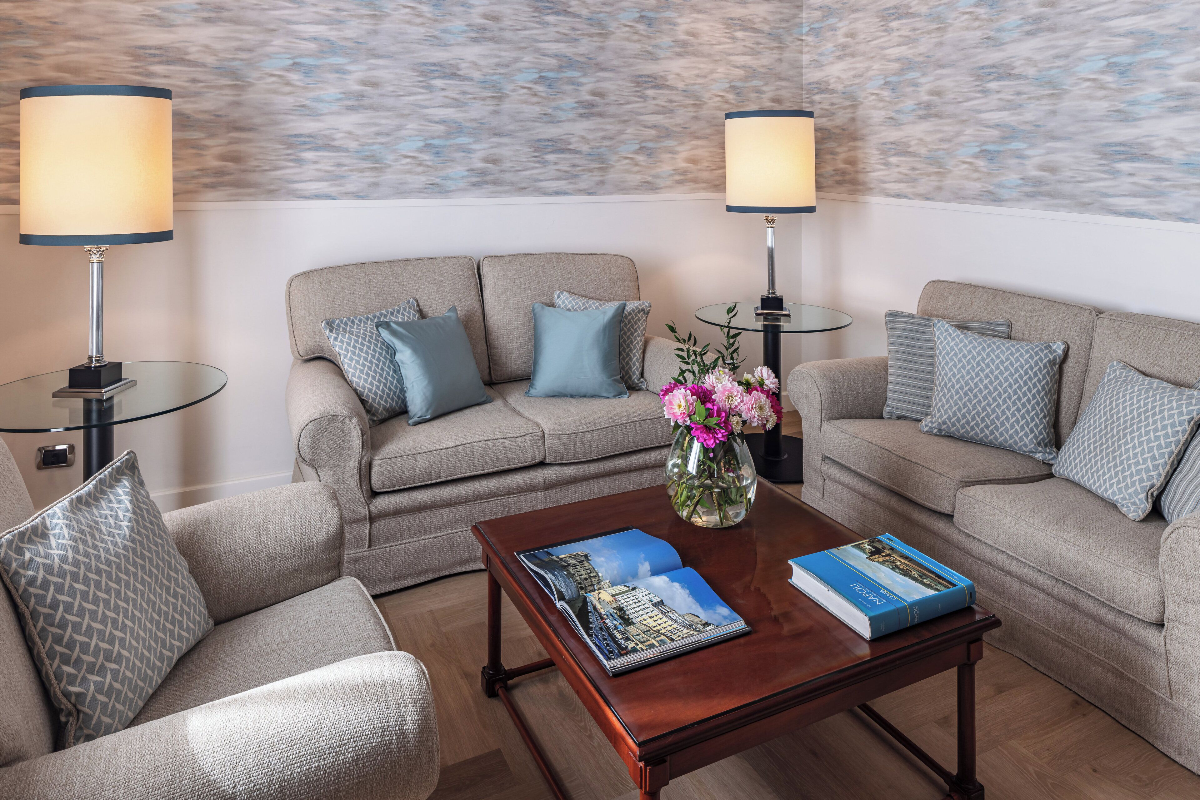 Una sala de estar moderna con un sofá beige, dos mesitas de noche con lámparas, una mesa de centro con un libro y flores, y una pared con textura.