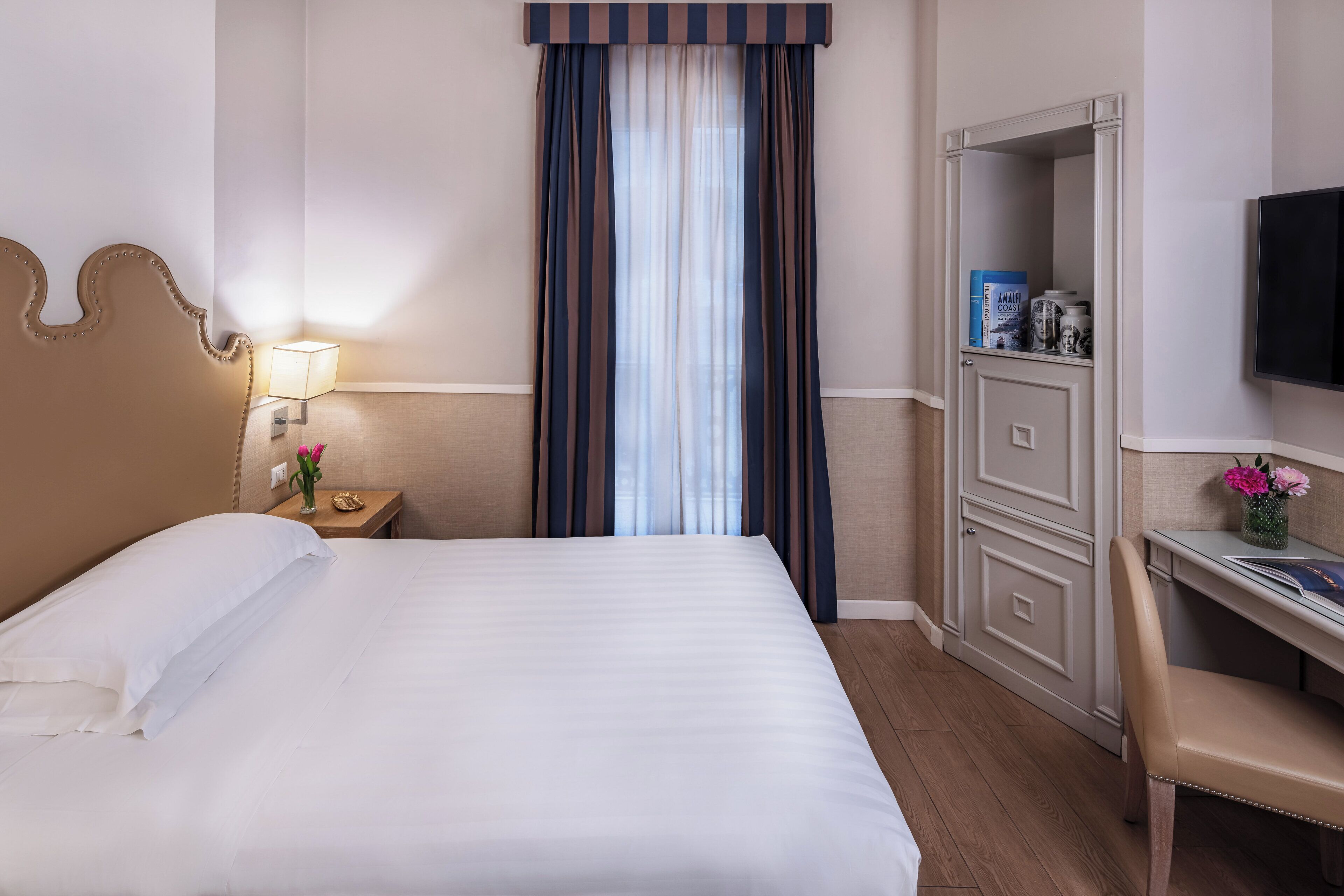 Een hotelkamer met een bed, een bureau, een stoel, een televisie, een nachtkastje en een raam met gordijnen.