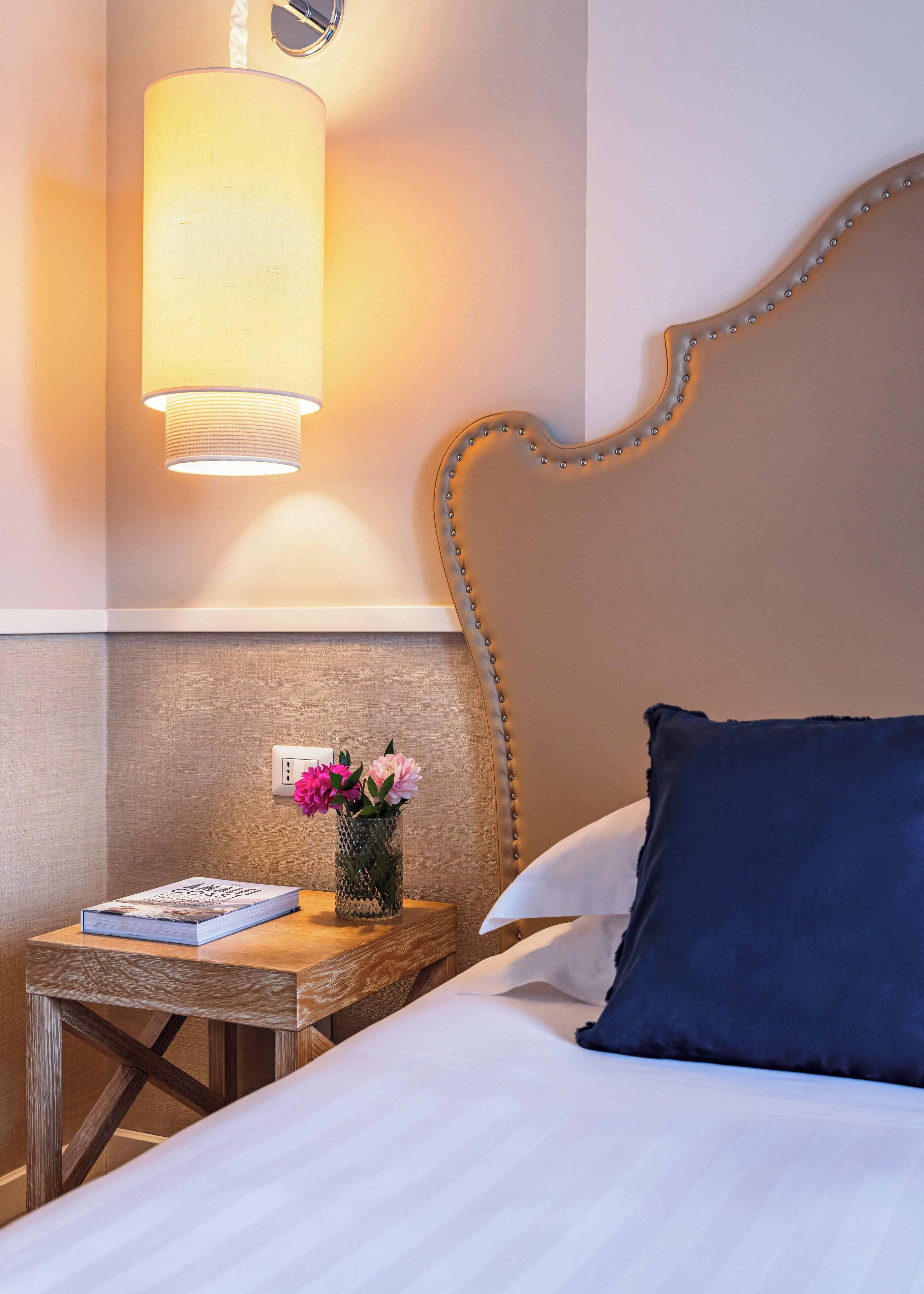 Ein ordentlich bezogenes Bett mit einem blauen Kopfkissen, ein Nachttisch aus Holz mit einem Buch und einer Blumenvase, sowie eine Wandlampe.