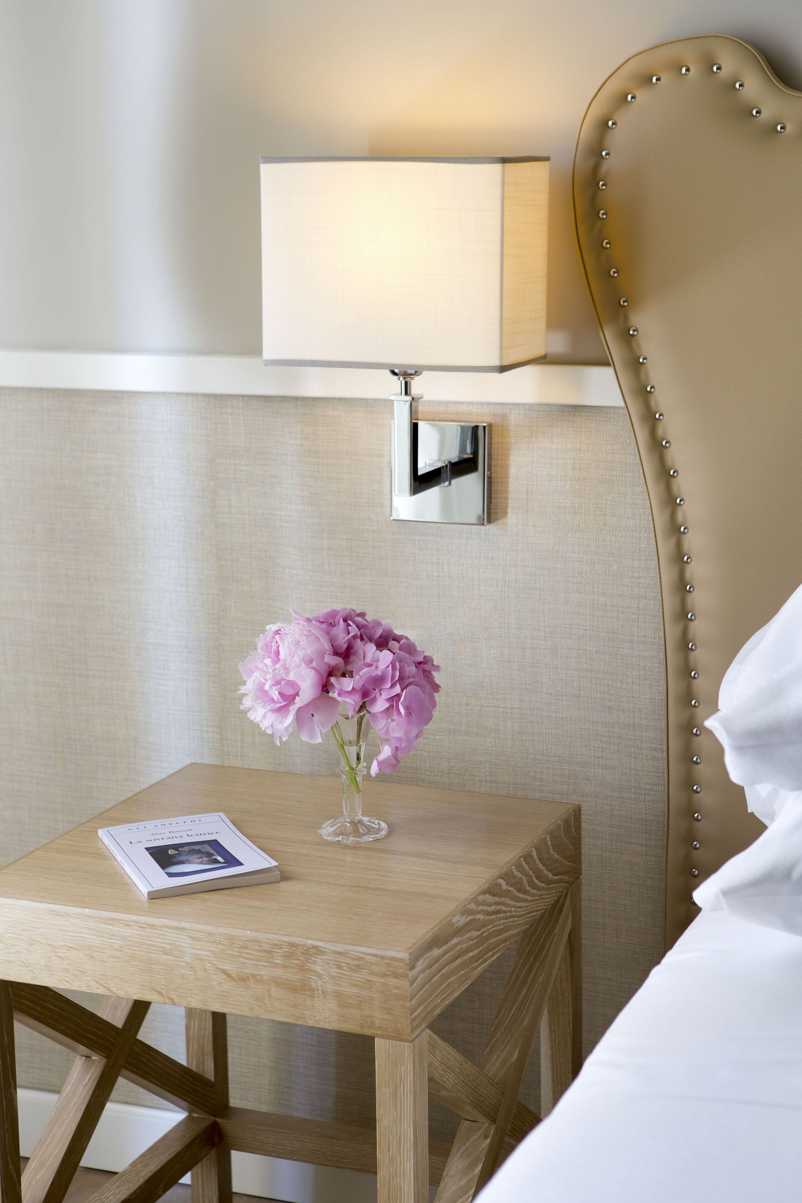 A neatly arranged hotel room with a bedside table, a lamp, a vase with flowers, and a book.