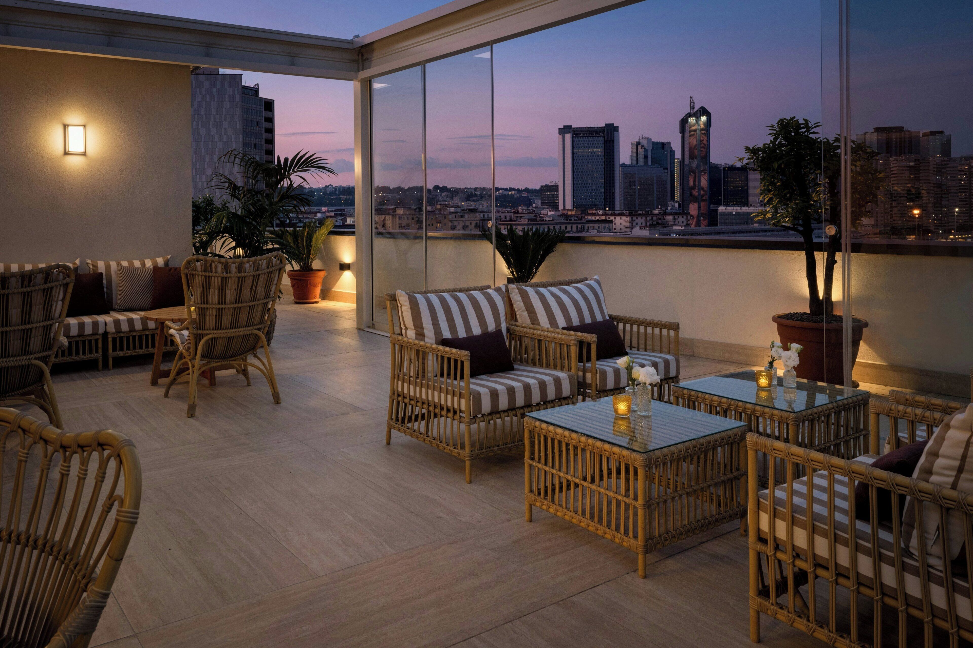 A rooftop terrace with wicker furniture, potted plants, and a city view at dusk.