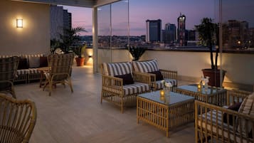 A rooftop terrace with wicker furniture, potted plants, and a city view at dusk.