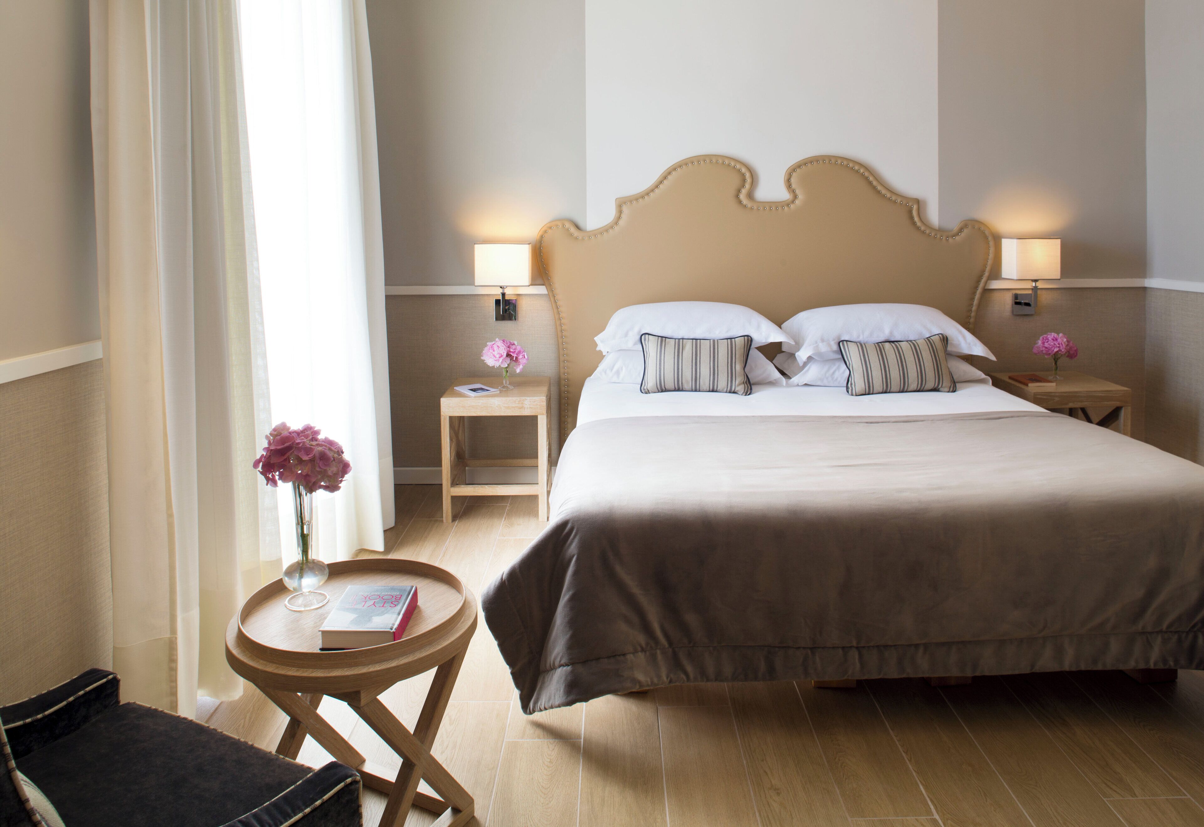 A hotel room with a large bed, two bedside tables with lamps, a small round table with a book and a vase of flowers, and a chair.