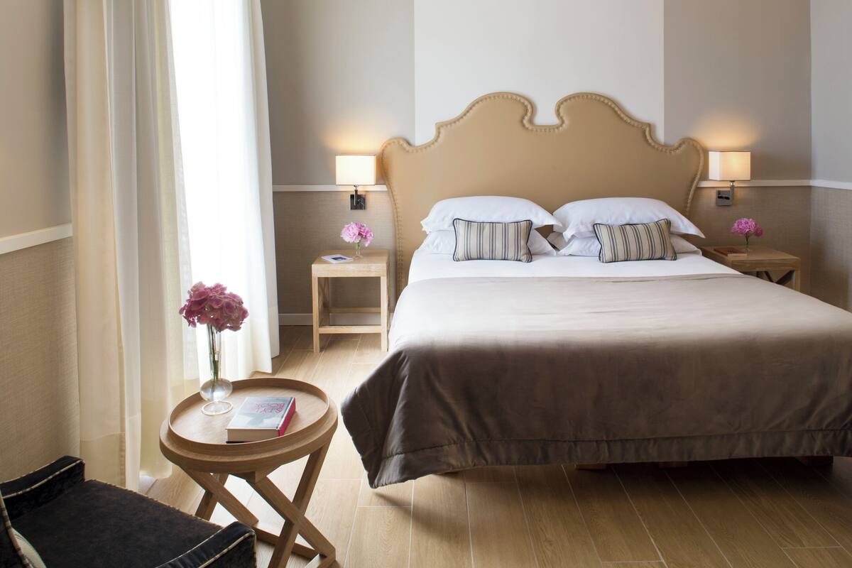 A hotel room with a large bed, two bedside tables with lamps, a small round table with a book and a vase of flowers, and a chair.