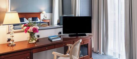 Una habitación de hotel con un escritorio de madera, una televisión, una silla, una cama con ropa de cama azul, un jarrón con flores y una ventana con cortinas.