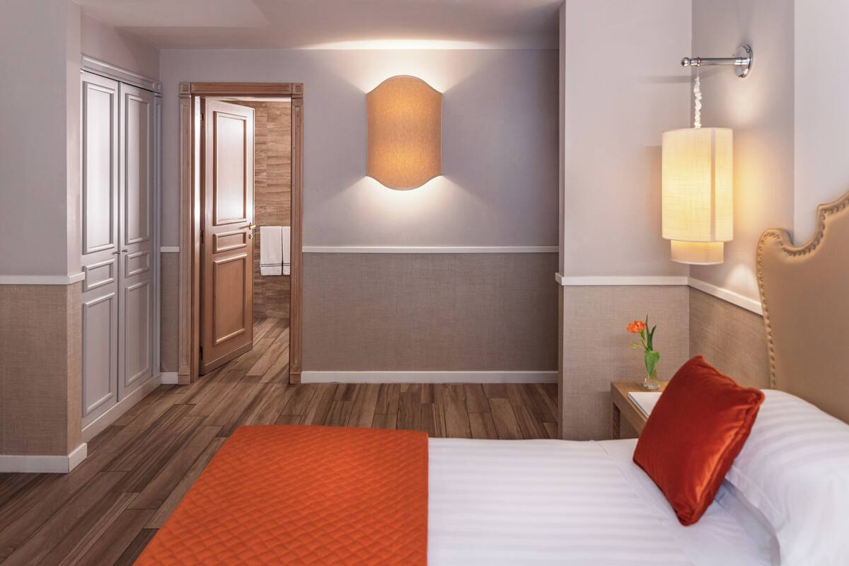A hotel room with a bed, a nightstand, a wall-mounted lamp, and a wooden door.
