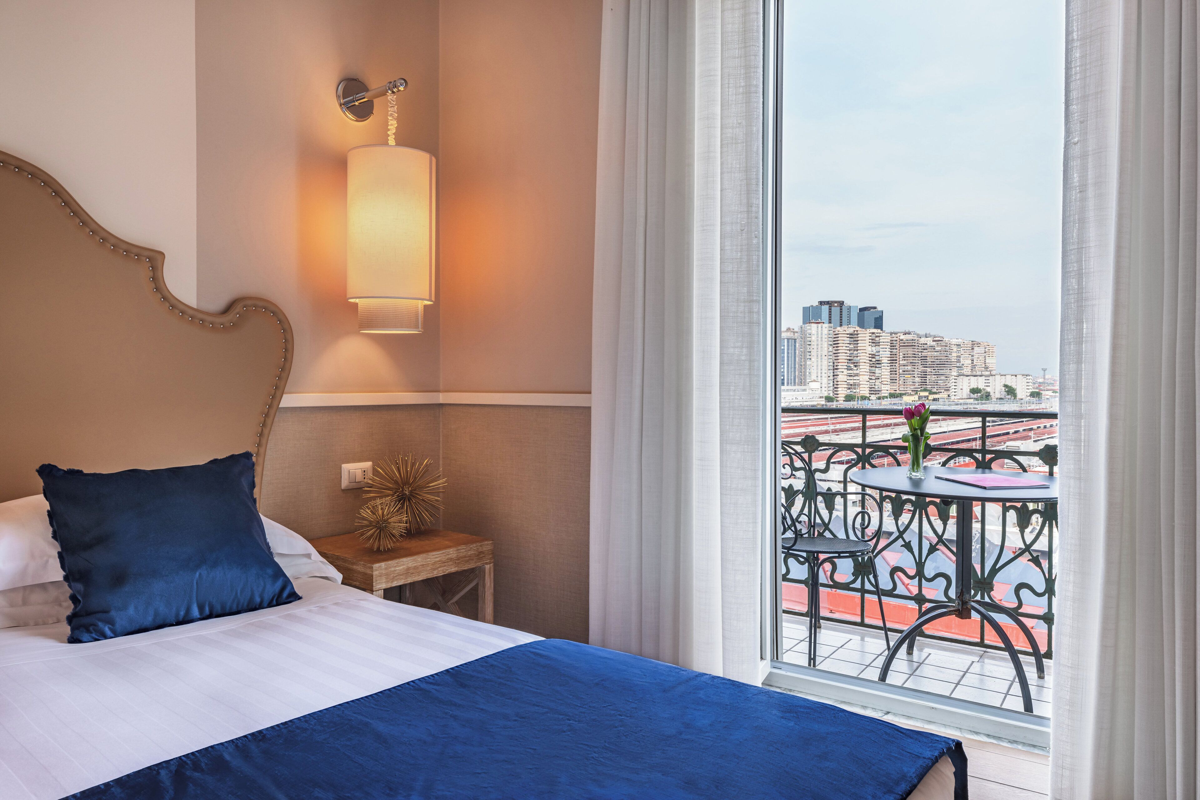 A hotel room with a large bed, a bedside table, a wall-mounted lamp, and a balcony with a view of the city.