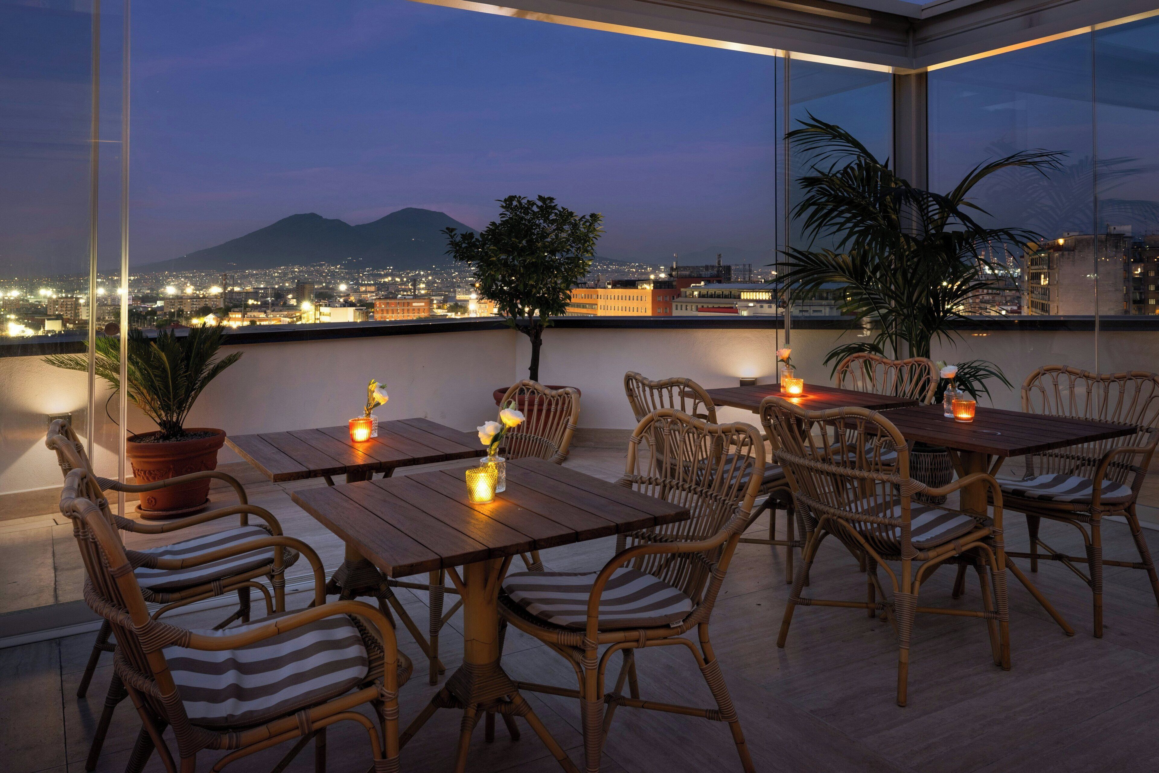 Una terrazza sul tetto con tavoli e sedie di legno, piante in vaso e vista sulla città al crepuscolo.