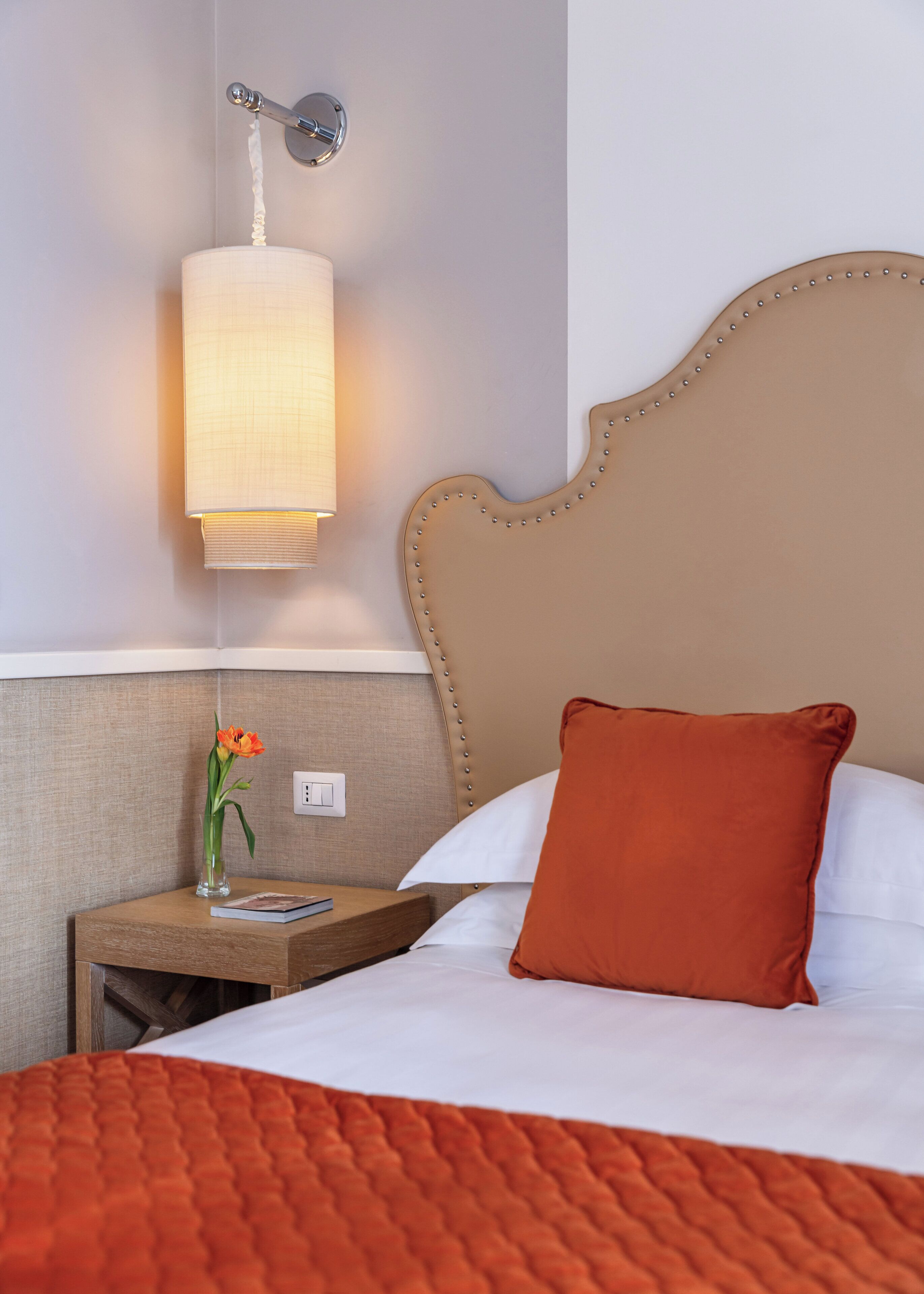A neatly made bed with an orange quilt, a wooden nightstand with a vase of flowers, and a wall-mounted lamp.