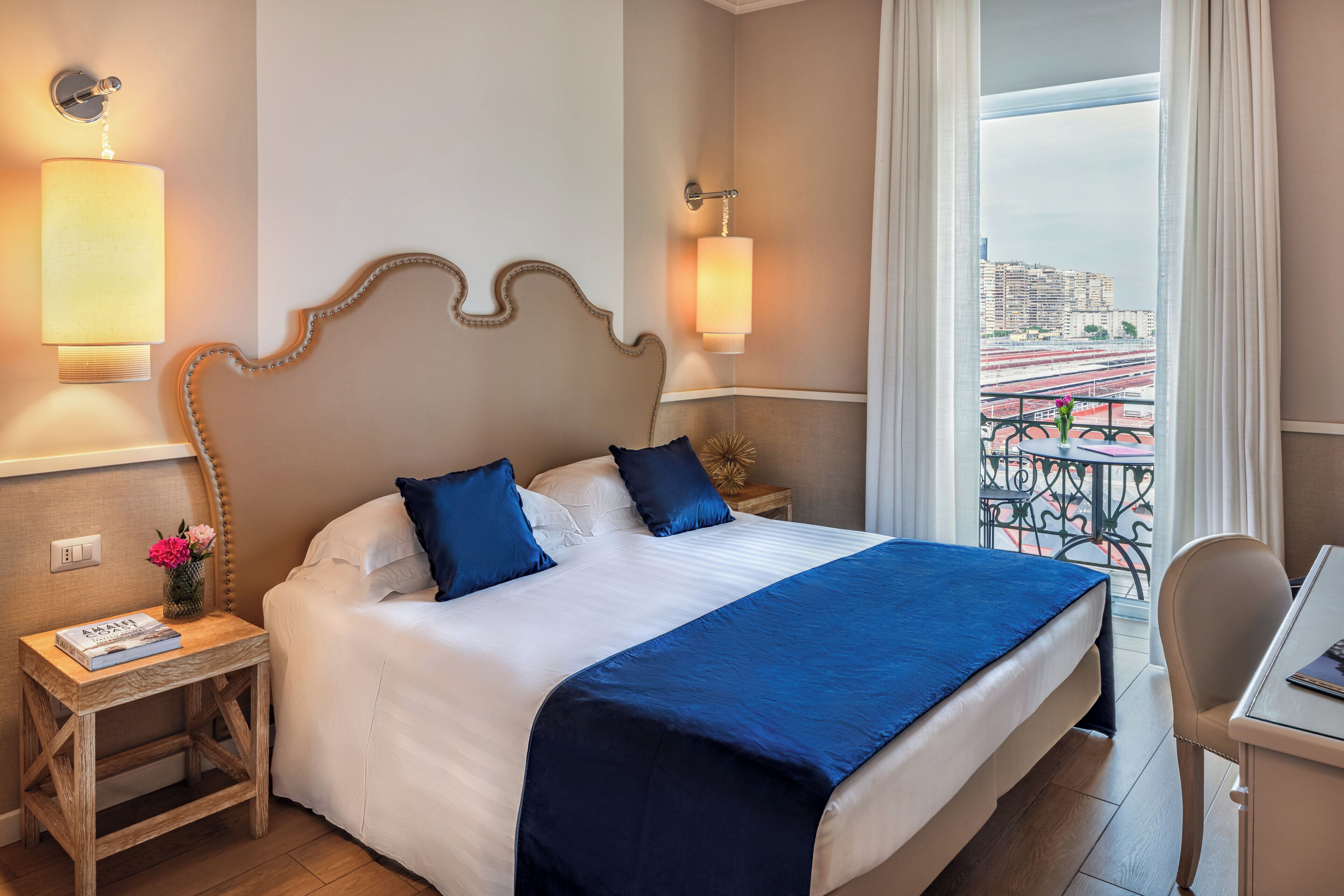 A hotel room with a large bed, two bedside lamps, a small table with a book, and a view of a cityscape through a balcony.