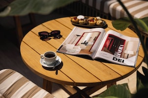 A round wooden table with a magazine, a cup of coffee, a pair of sunglasses, and a plate of pastries.