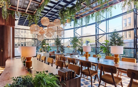 A spacious indoor area with a long wooden table, multiple chairs, hanging wicker lamps, and potted plants.