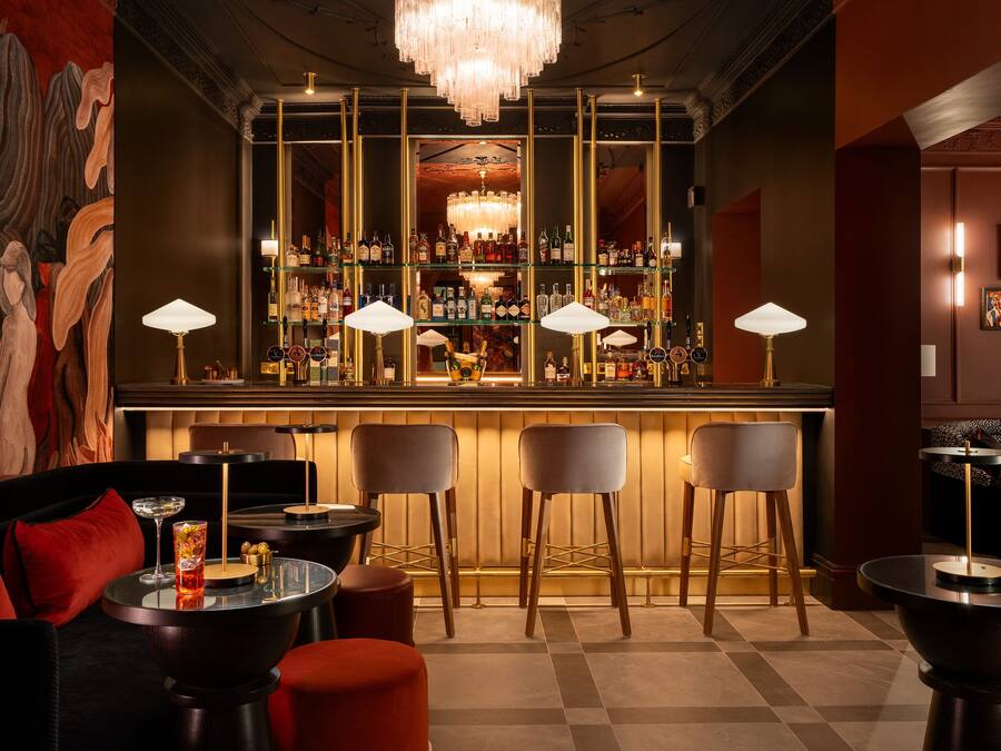 A bar area with a chandelier, multiple bar stools, and a variety of bottles on shelves.