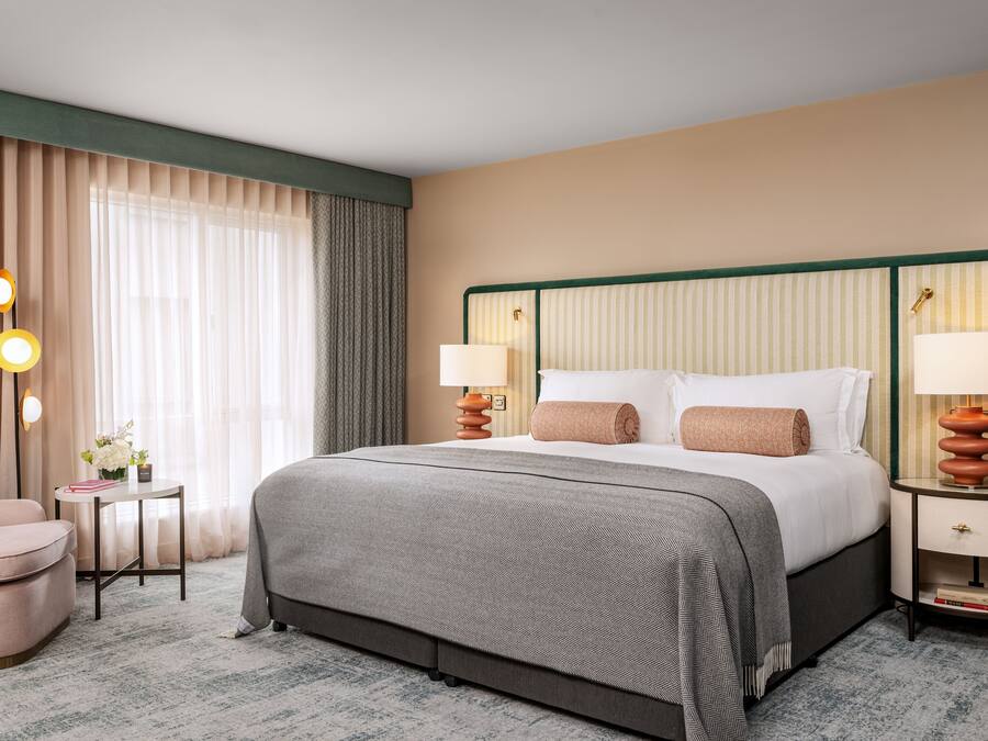 Quarto de hotel com uma cama grande, duas mesas de cabeceira com luminárias, uma mesinha lateral, uma cadeira e uma janela ampla com cortinas.