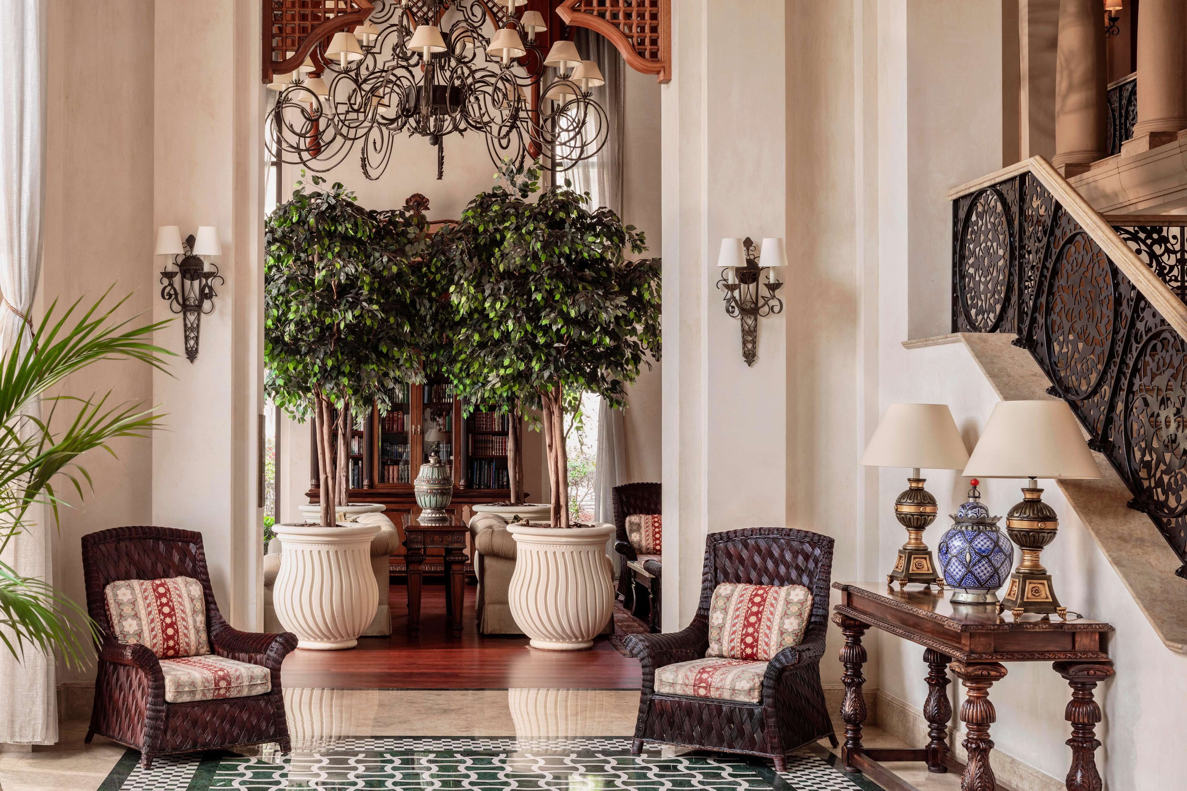 A grand lobby with a large potted plant, two armchairs, a wooden table with lamps, and a staircase with ornate railings.