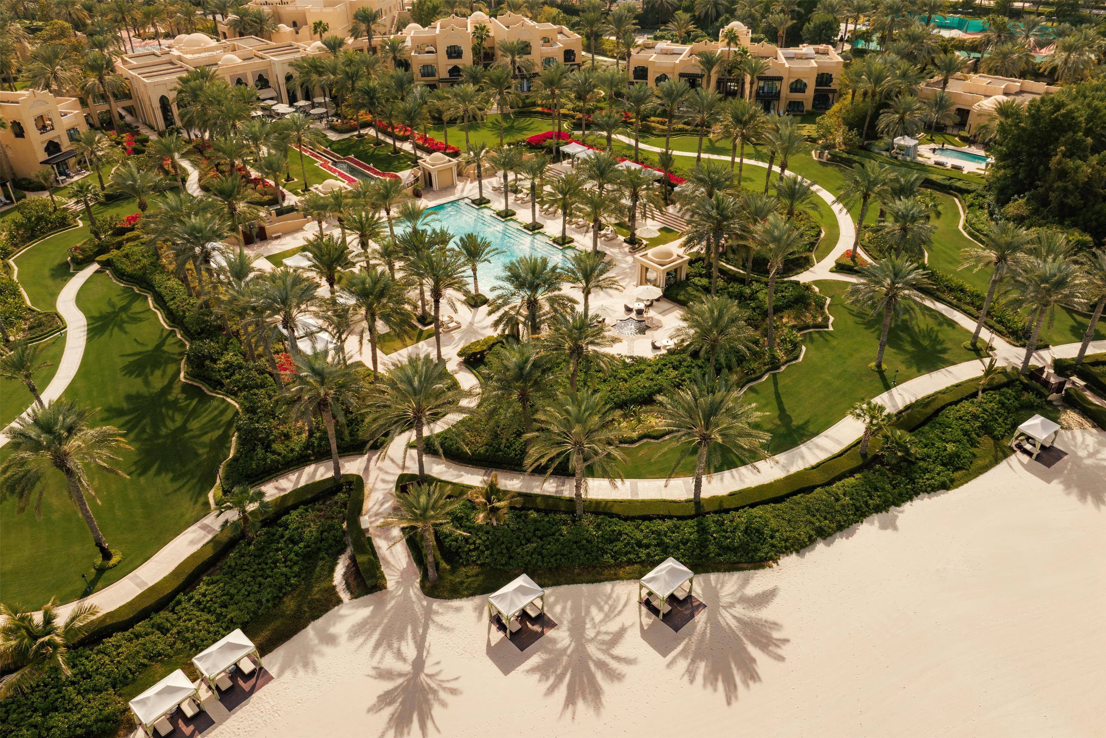 An aerial view of a resort with a central pool, palm trees, and a beach area with sun loungers.