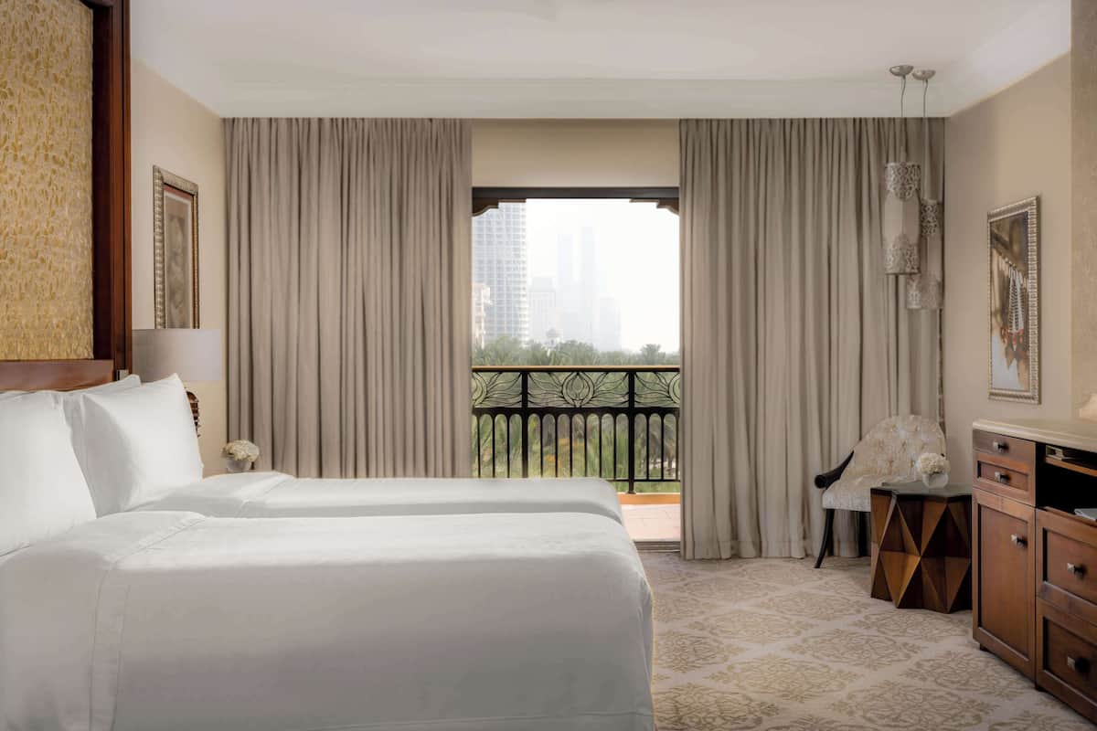 Ein Hotelzimmer mit zwei Betten, Blick auf die Stadt durch große Fenster, einem Balkon und einem kleinen Beistelltisch mit einem Sessel.