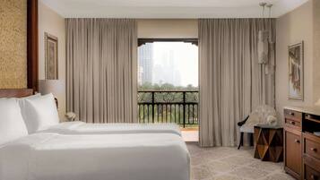 A hotel room with two beds, a view of the city through large windows, a balcony, and a small side table with a chair.