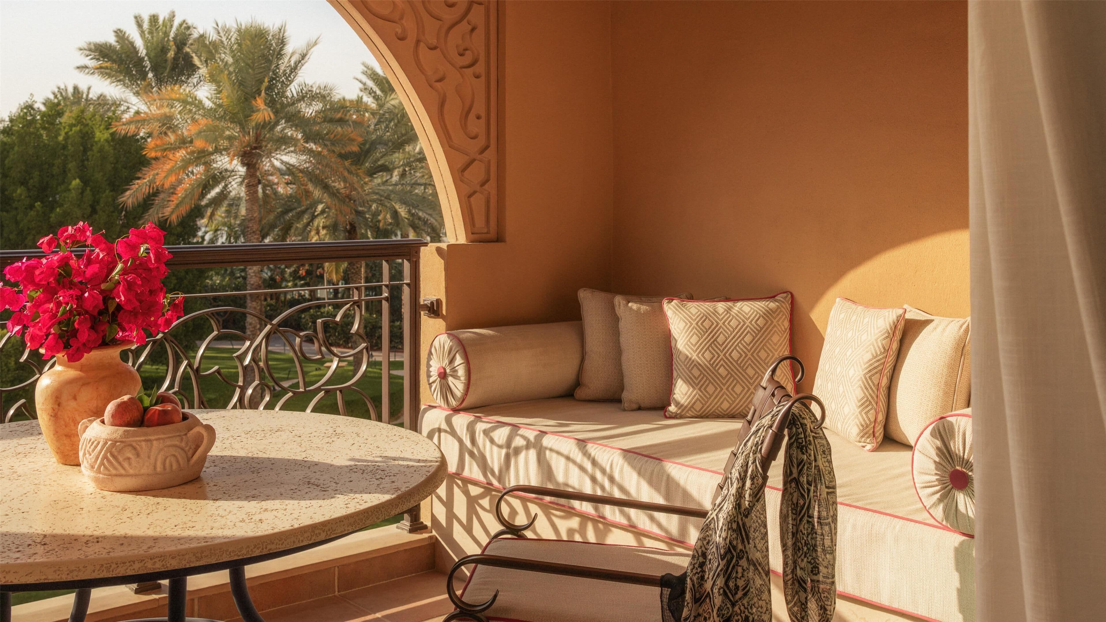 A balcony with a round table, a sofa with patterned cushions, and a vase of red flowers.