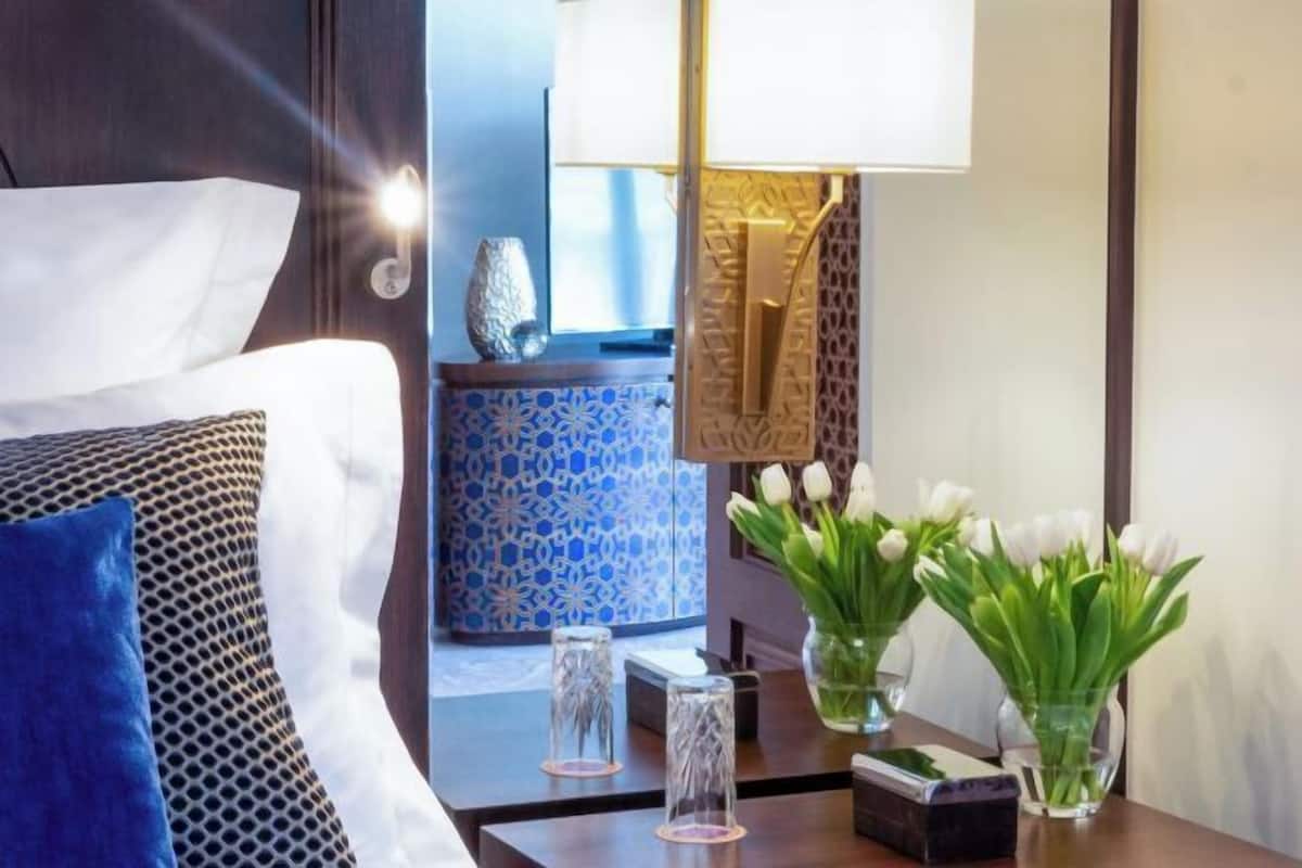 Ein Hotelzimmer mit Bett, Nachttisch mit Lampe, einer dekorativen Vase mit Blumen und einer blauen Wand mit Muster.