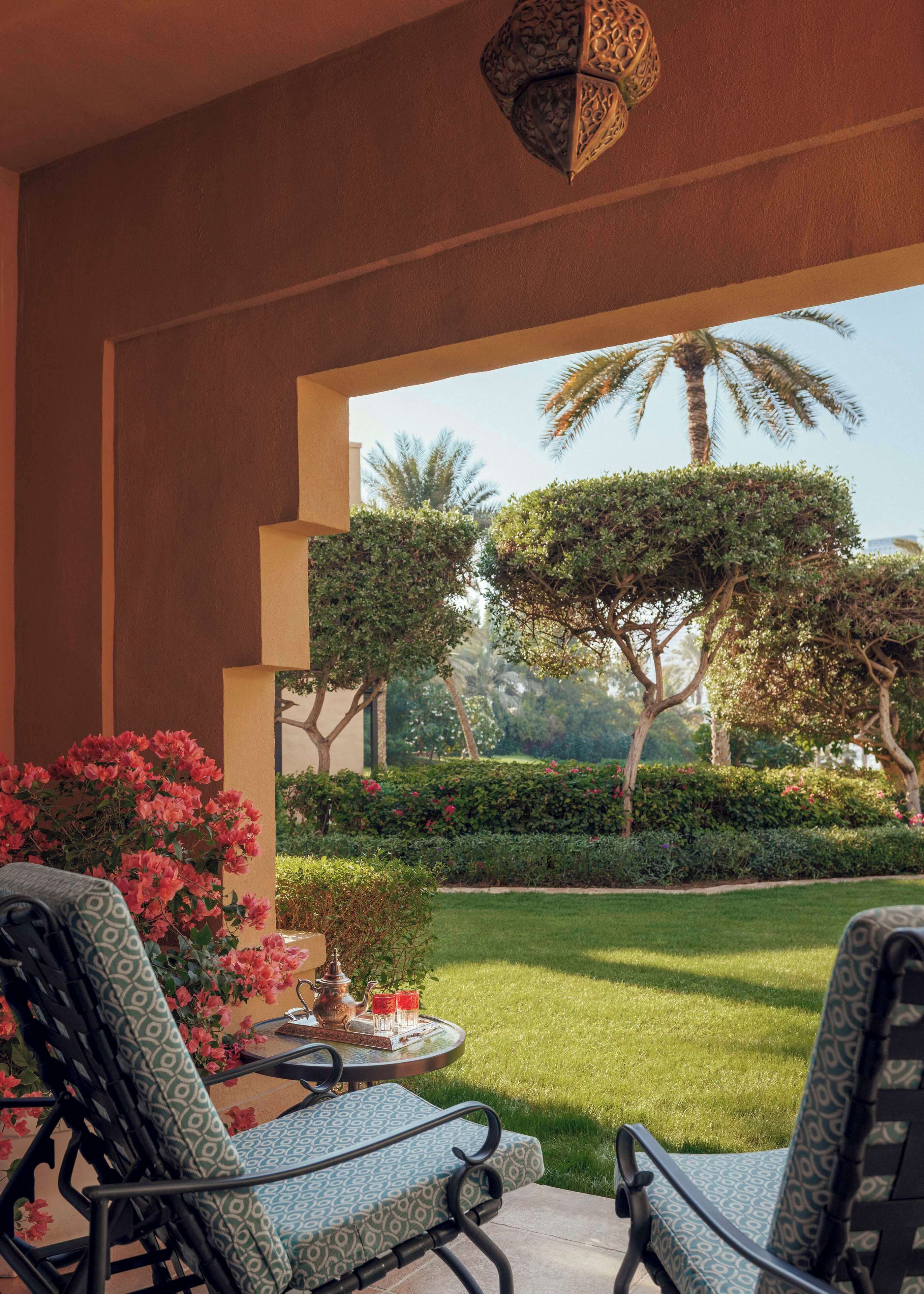 A patio with two patterned chairs, a small table with a teapot and glasses, and a view of a well-manicured garden with palm trees and shrubs.