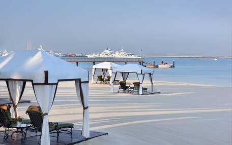 A beachside area with white canopies and lounge chairs, a pier, and a large yacht in the background.