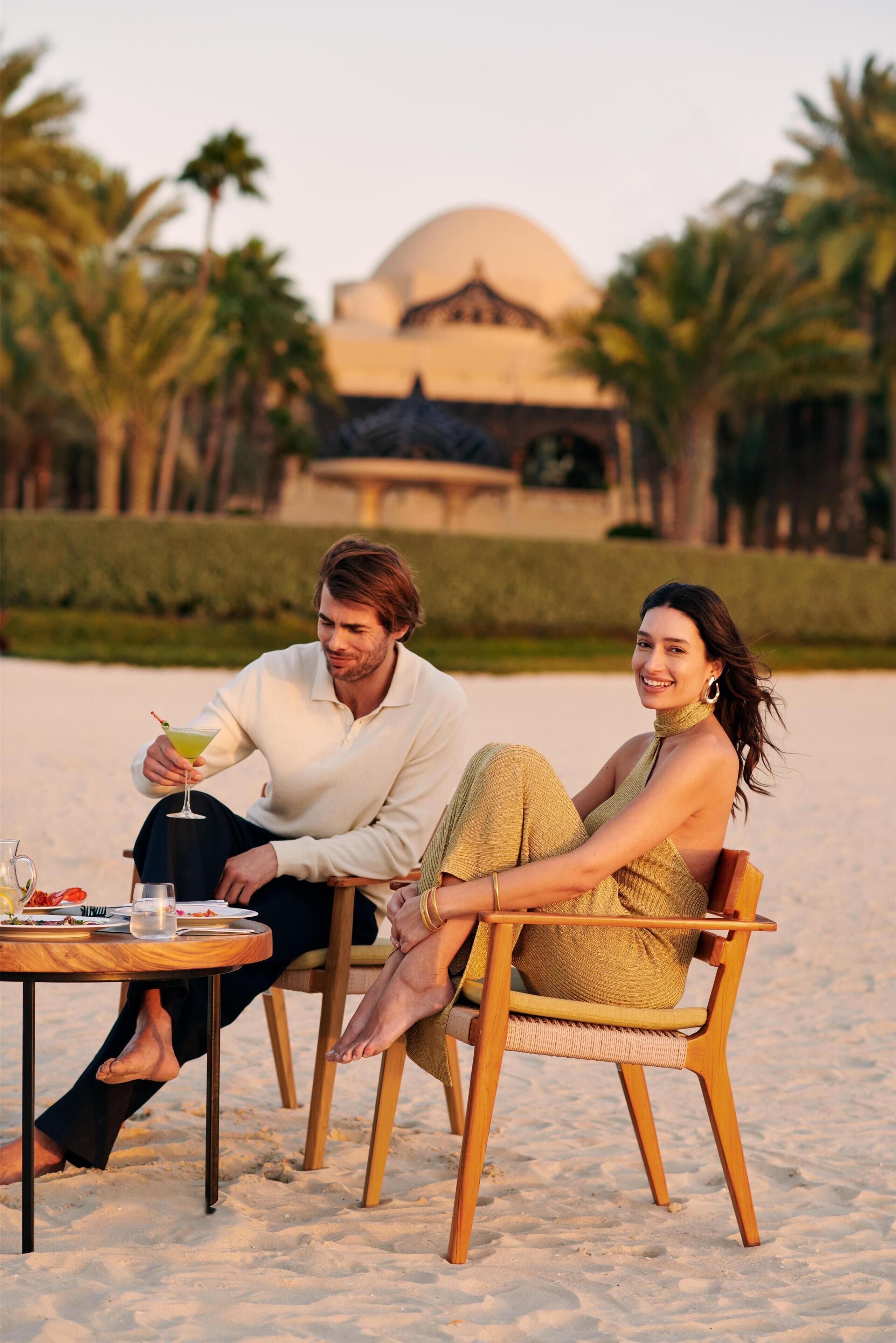Two people sitting on a wooden chair on a sandy beach with a table in front of them.