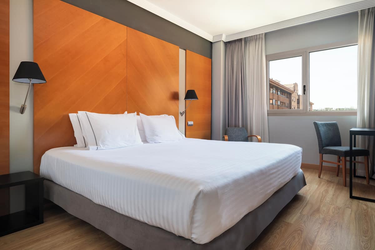 A hotel room with a large bed, two bedside tables, a chair, a desk, and a window with curtains.