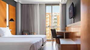 Habitación de hotel con cama, escritorio, silla, televisión y ventana con vistas a edificios.