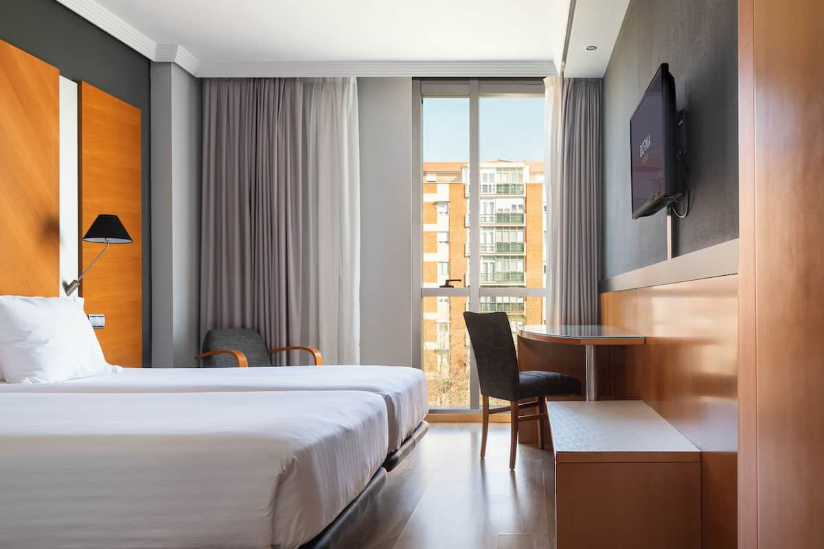 A hotel room with a bed, a desk, a chair, a TV, and a window with a view of buildings.