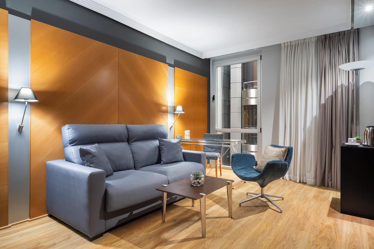 A modern living room with a grey sofa, a small table, a blue chair, a lamp, and a kitchen area visible through a glass partition.
