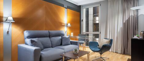 Una sala de estar moderna con un sofá gris, una mesita, un sillón azul, una lámpara y una cocina visible a través de un separador de vidrio.