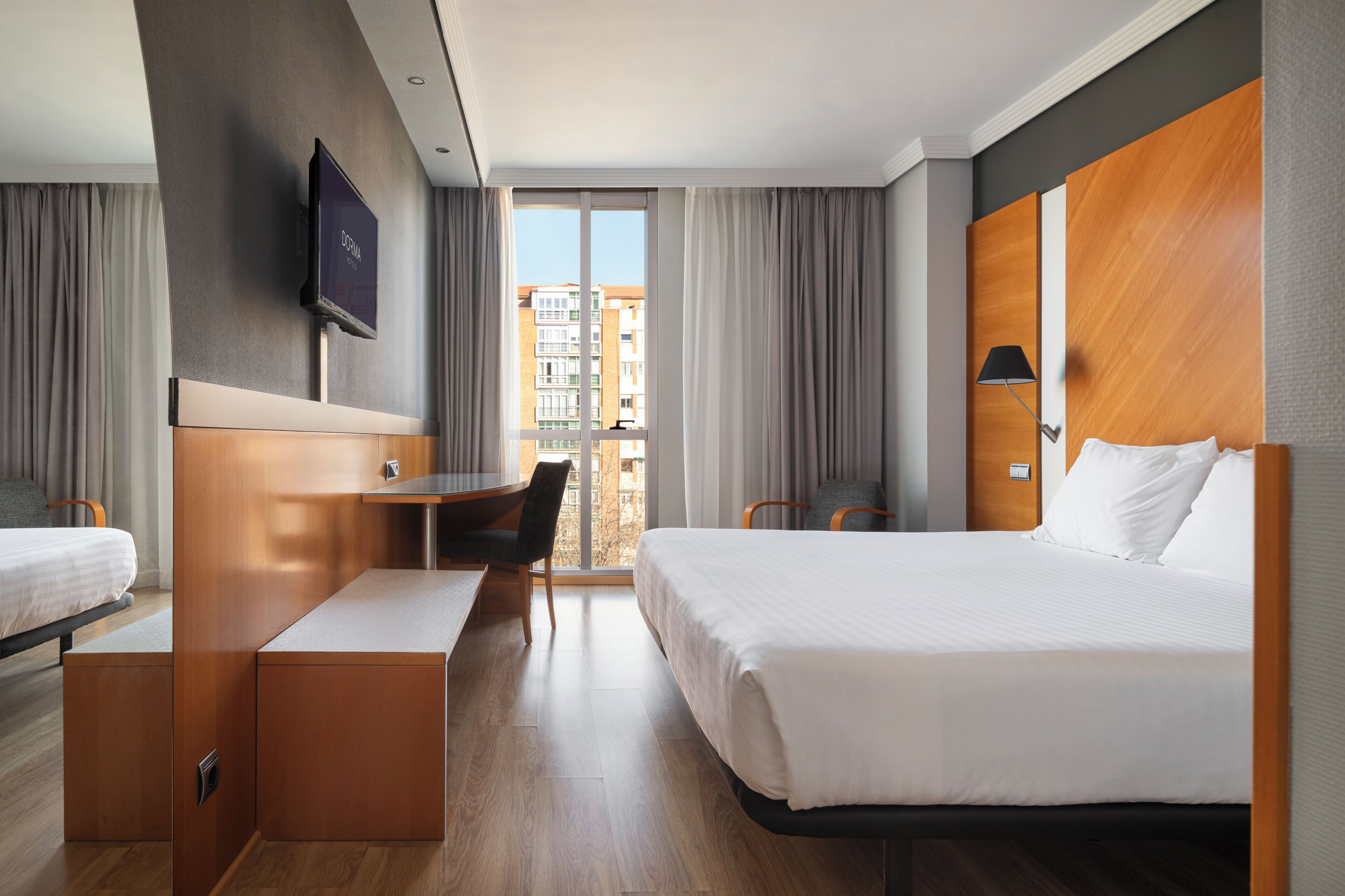 A hotel room with a bed, a desk, a chair, a TV, and a view of buildings through the window.