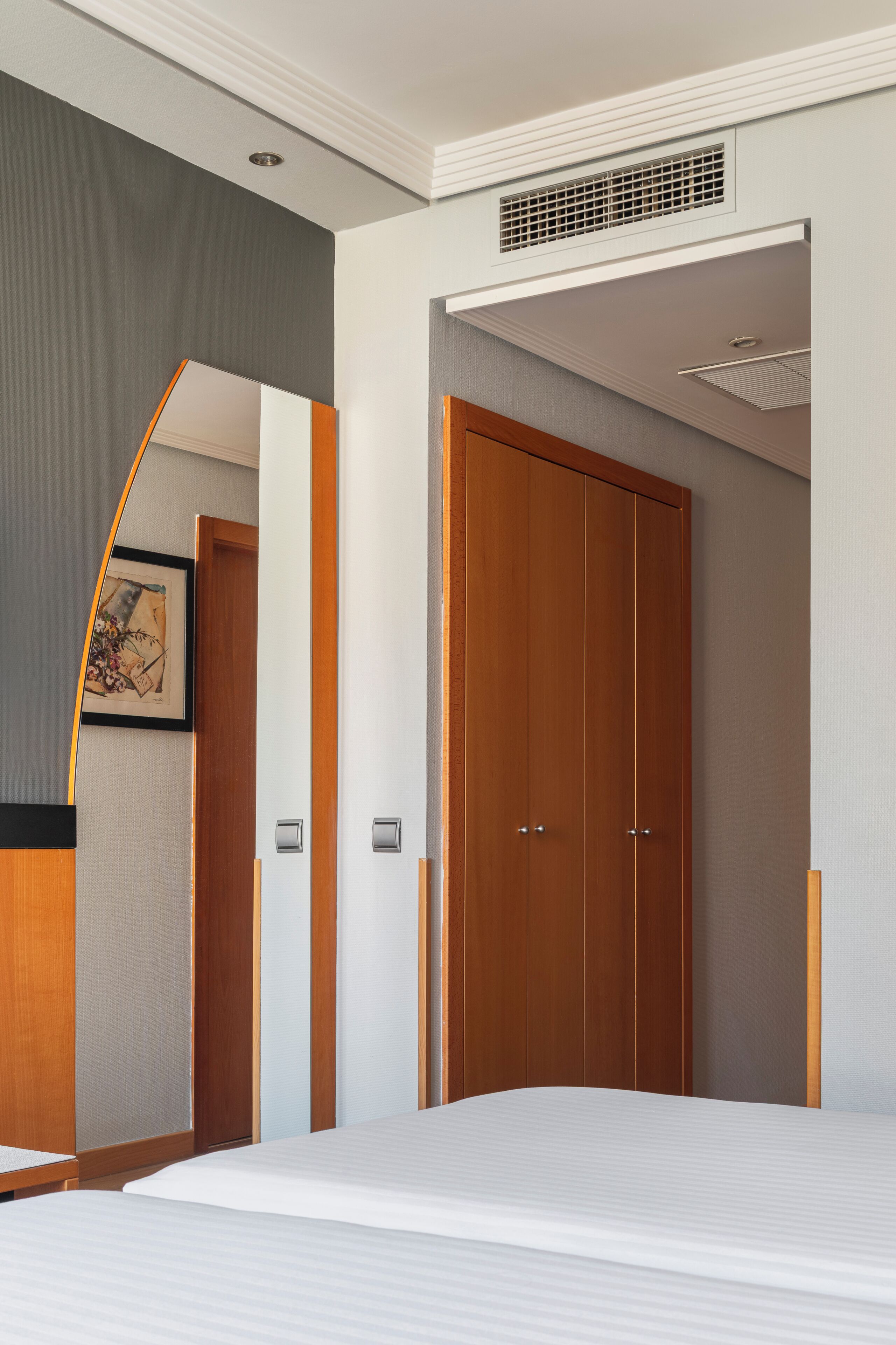 Habitación de hotel con cama, armario de madera, un cuadro enmarcado en la pared y una rejilla en el techo.
